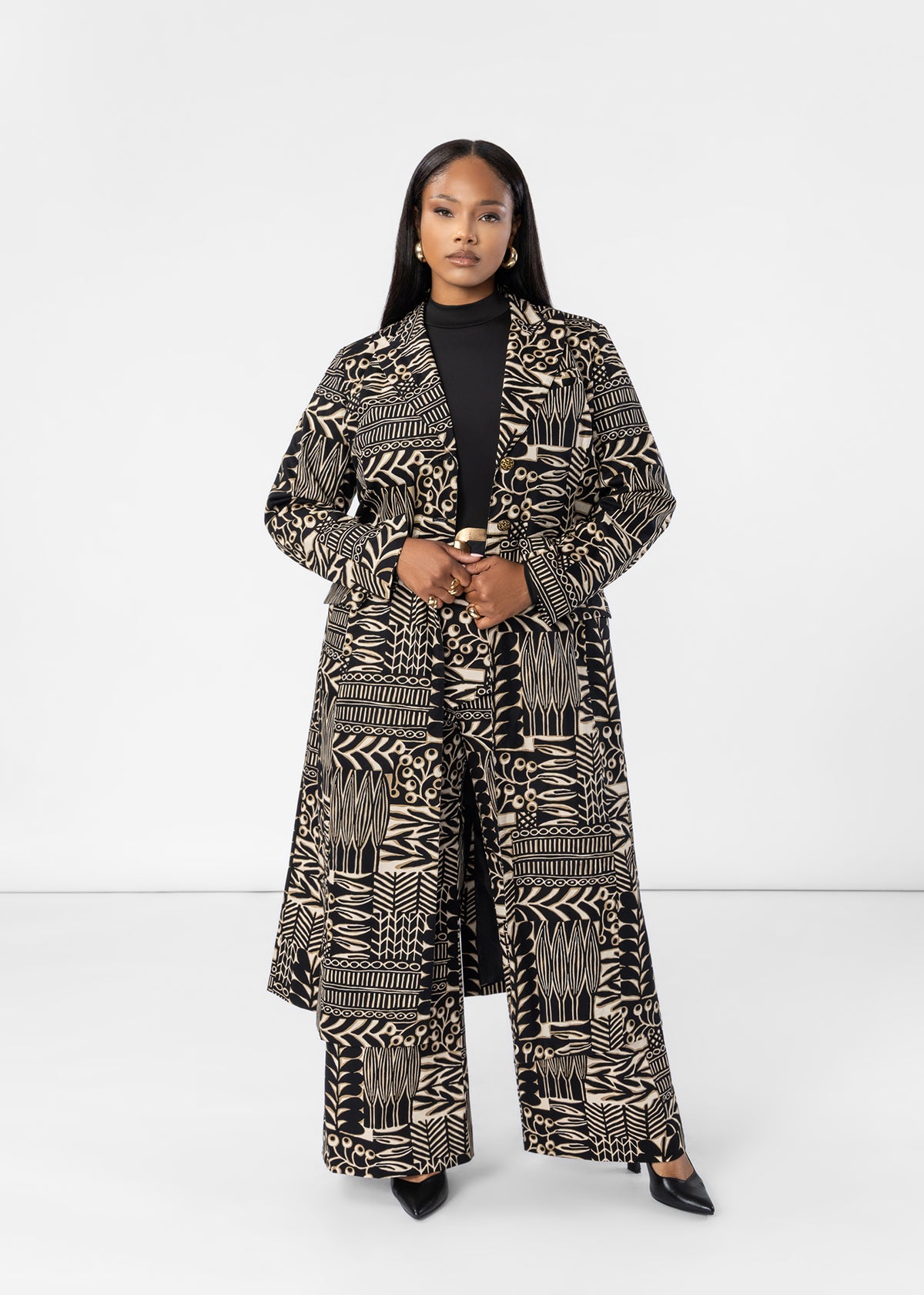 A woman modeling a long black and beige geometric patterned coat over a black top and wide-leg pants, standing against a plain light gray studio background.
