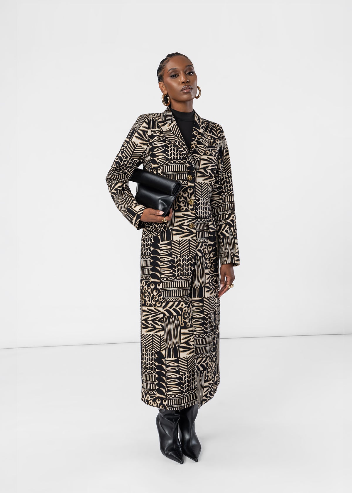 A model stands in a studio wearing a long black and beige geometric patterned coat over a black top, holding a black clutch bag and wearing black pointed boots.