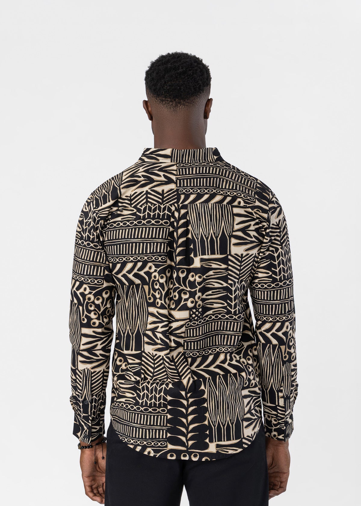A man viewed from the back wearing a long-sleeve button-up shirt with an intricate black and beige tribal-style geometric pattern, paired with black trousers, against a plain light gray background.