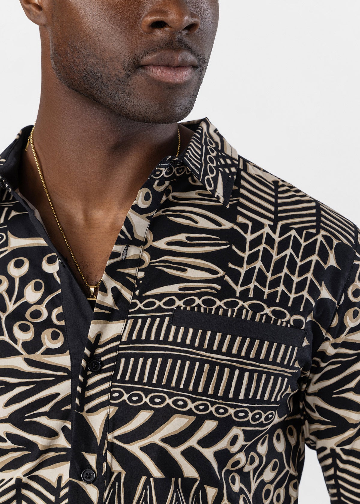 A close-up of a man wearing a black and beige patterned button-down shirt with a collar and chest pocket, paired with a gold chain necklace, against a light gray background.