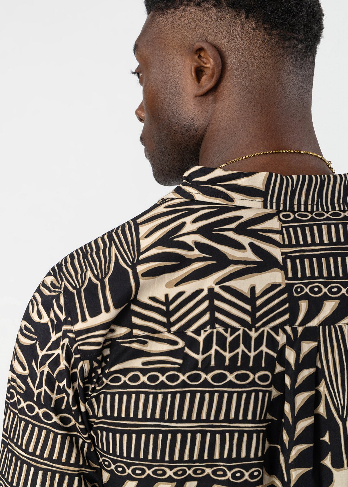 A close-up side profile of a man wearing a black and beige patterned shirt with geometric and leaf-like motifs, set against a plain light gray background.