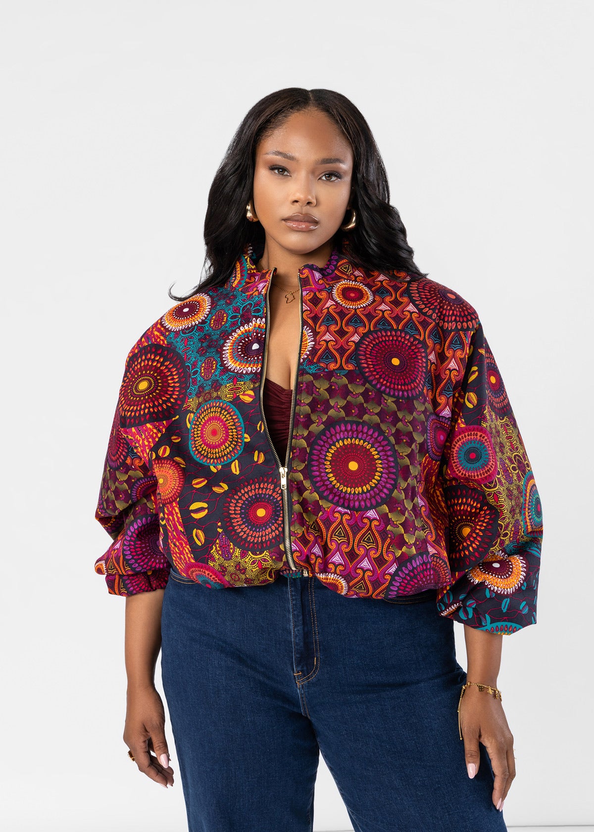 A woman modeling a vibrant, multicolored patterned bomber jacket with balloon sleeves, paired with dark blue jeans, against a plain white studio background.