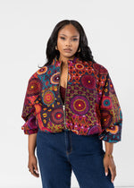 A woman modeling a vibrant, multicolored patterned bomber jacket with balloon sleeves, paired with dark blue jeans, against a plain white studio background.
