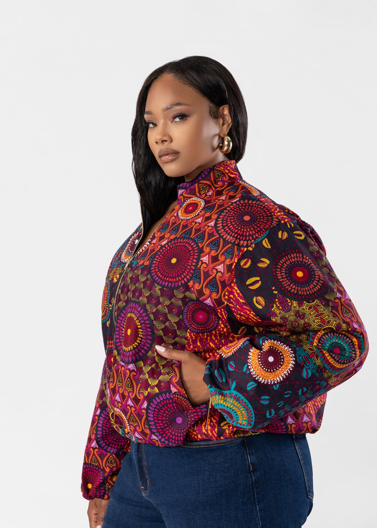 A woman modeling a cropped, long-sleeve jacket with a vibrant, multicolored African wax print pattern featuring circular mandala designs, paired with blue jeans against a light gray studio background.