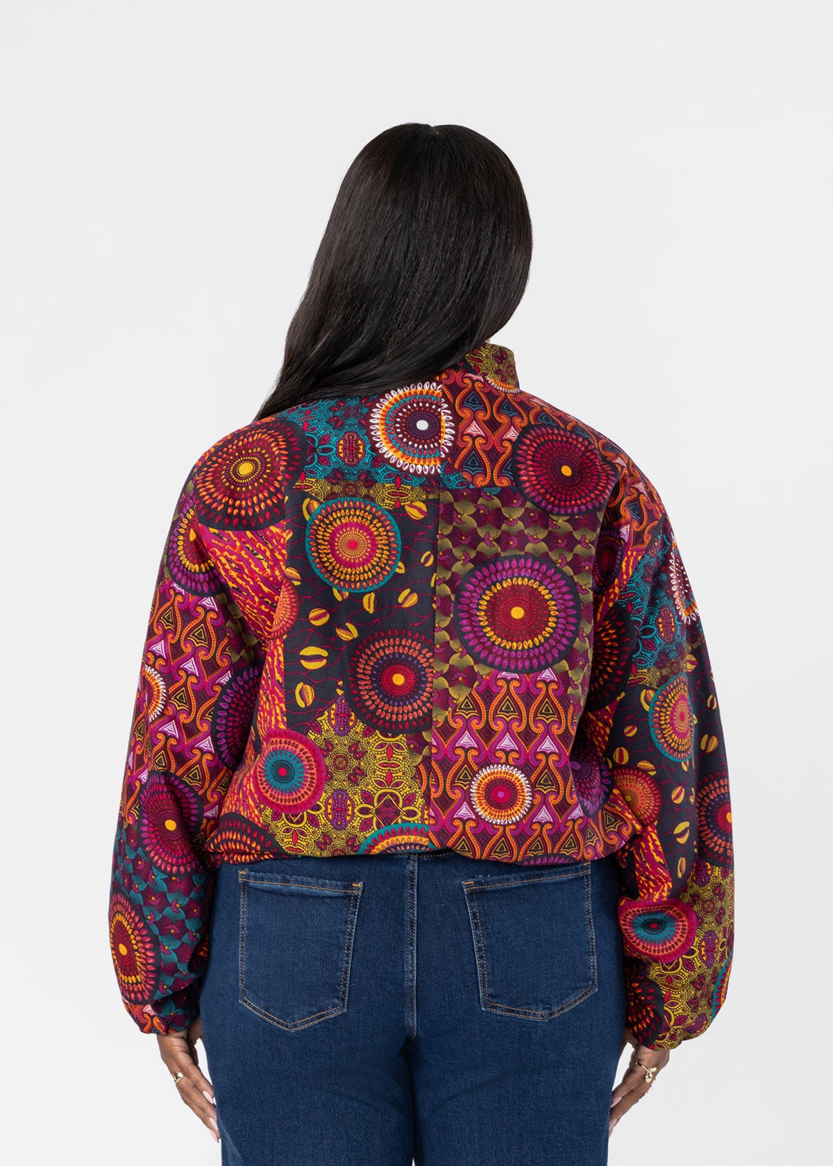 A woman viewed from the back wearing a colorful patchwork jacket with intricate circular and geometric patterns in red, orange, yellow, blue, and purple, paired with dark blue jeans against a plain white background.
