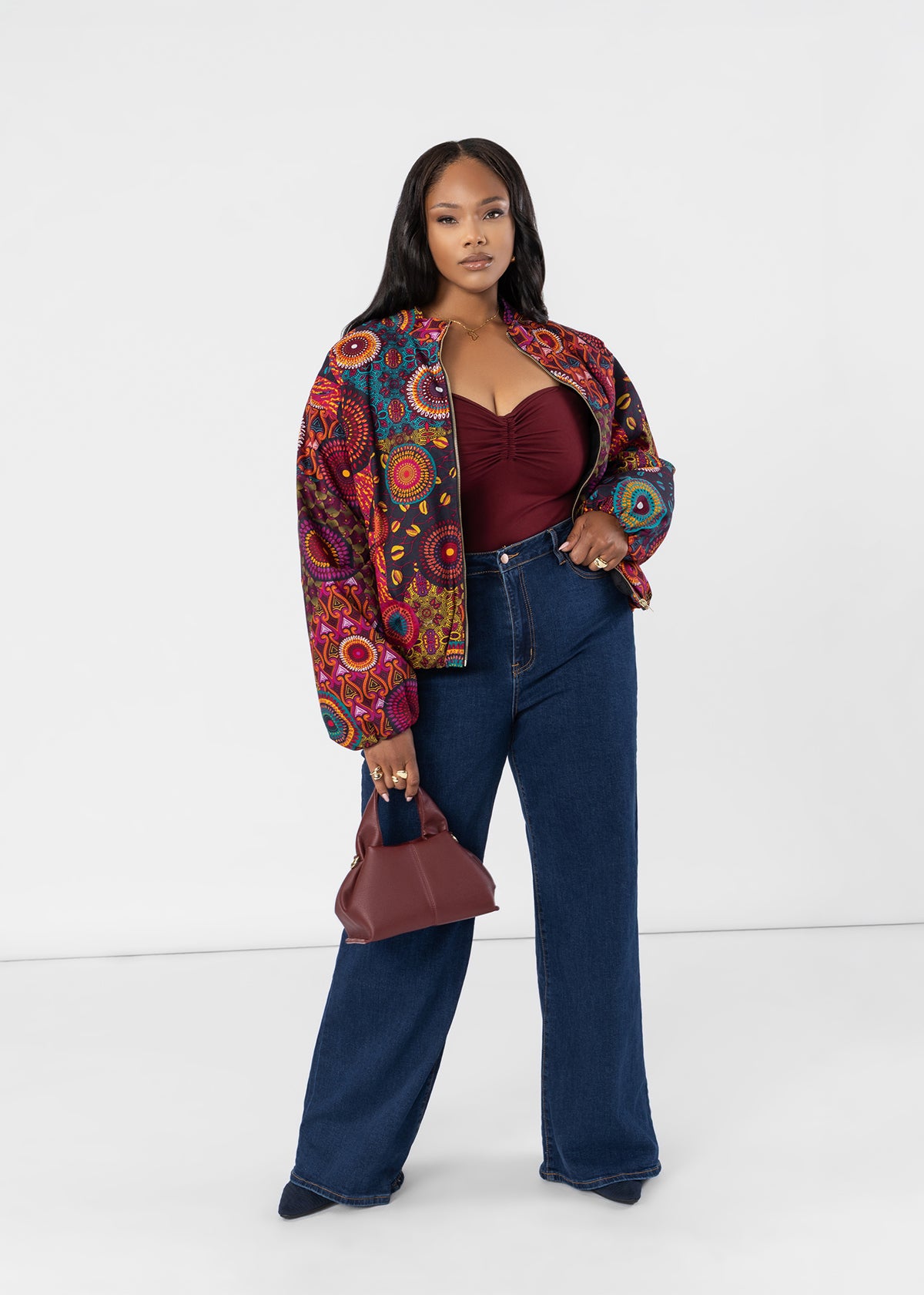 A woman stands against a clean white studio background wearing a colorful patterned bomber jacket over a dark red sweetheart top and dark blue wide-leg jeans, holding a small burgundy handbag.