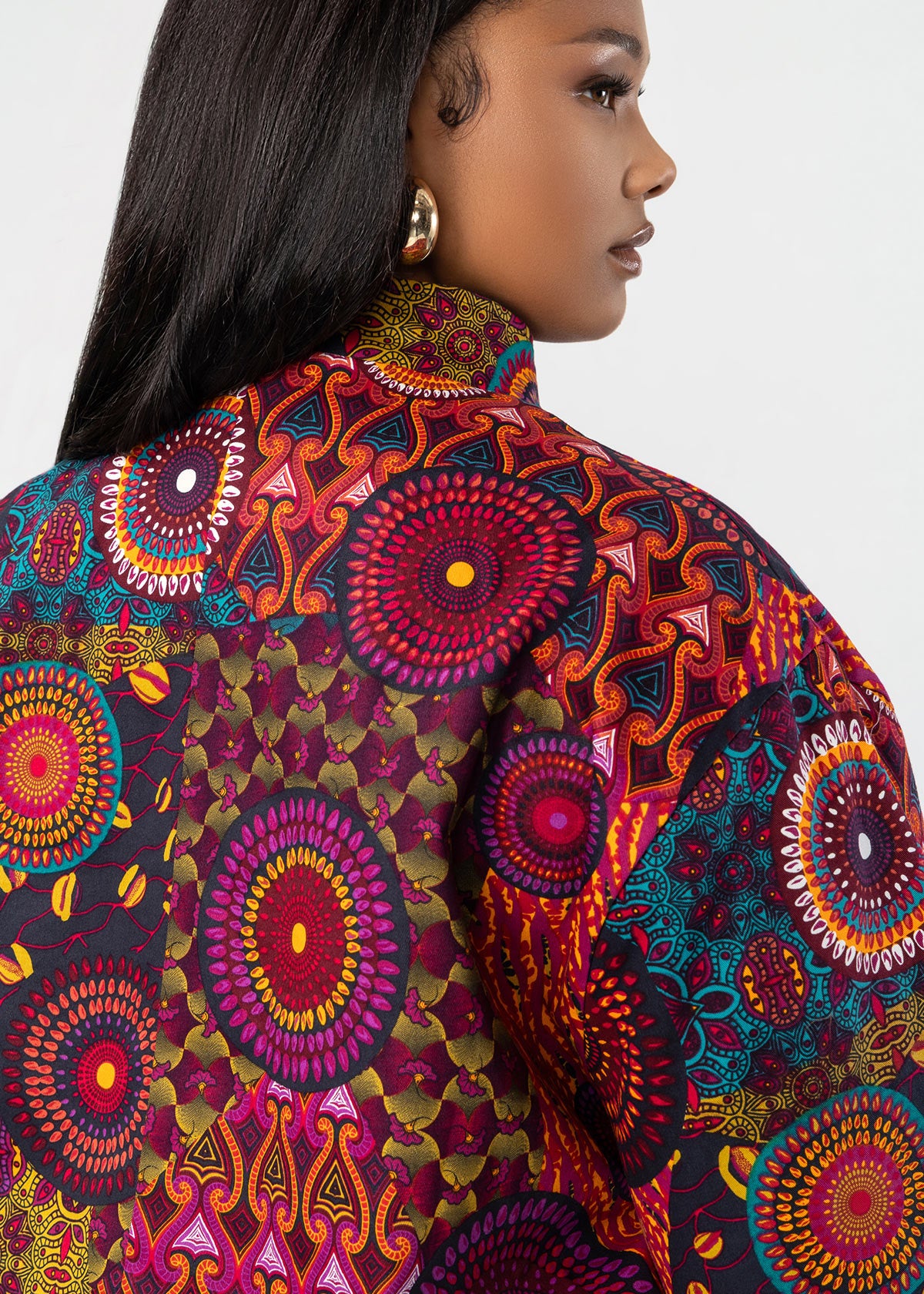 A close-up side profile of a model wearing a vibrant African wax print blouse with intricate circular and geometric patterns in red, purple, orange, and teal.