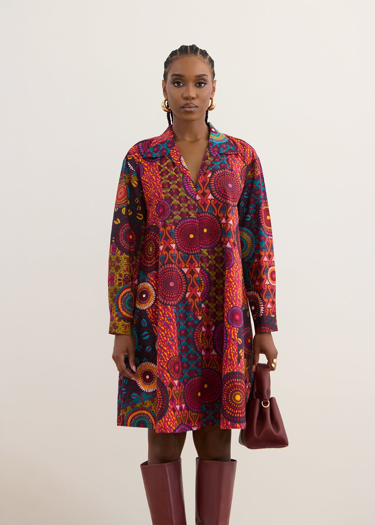 A model stands against a plain light beige background wearing a long-sleeve collared shirt dress with a vibrant, multicolored African wax print pattern. She is also wearing dark burgundy knee-high boots and holding a matching dark burgundy handbag.
