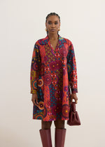 A model stands against a plain light beige background wearing a long-sleeve collared shirt dress with a vibrant, multicolored African wax print pattern. She is also wearing dark burgundy knee-high boots and holding a matching dark burgundy handbag.