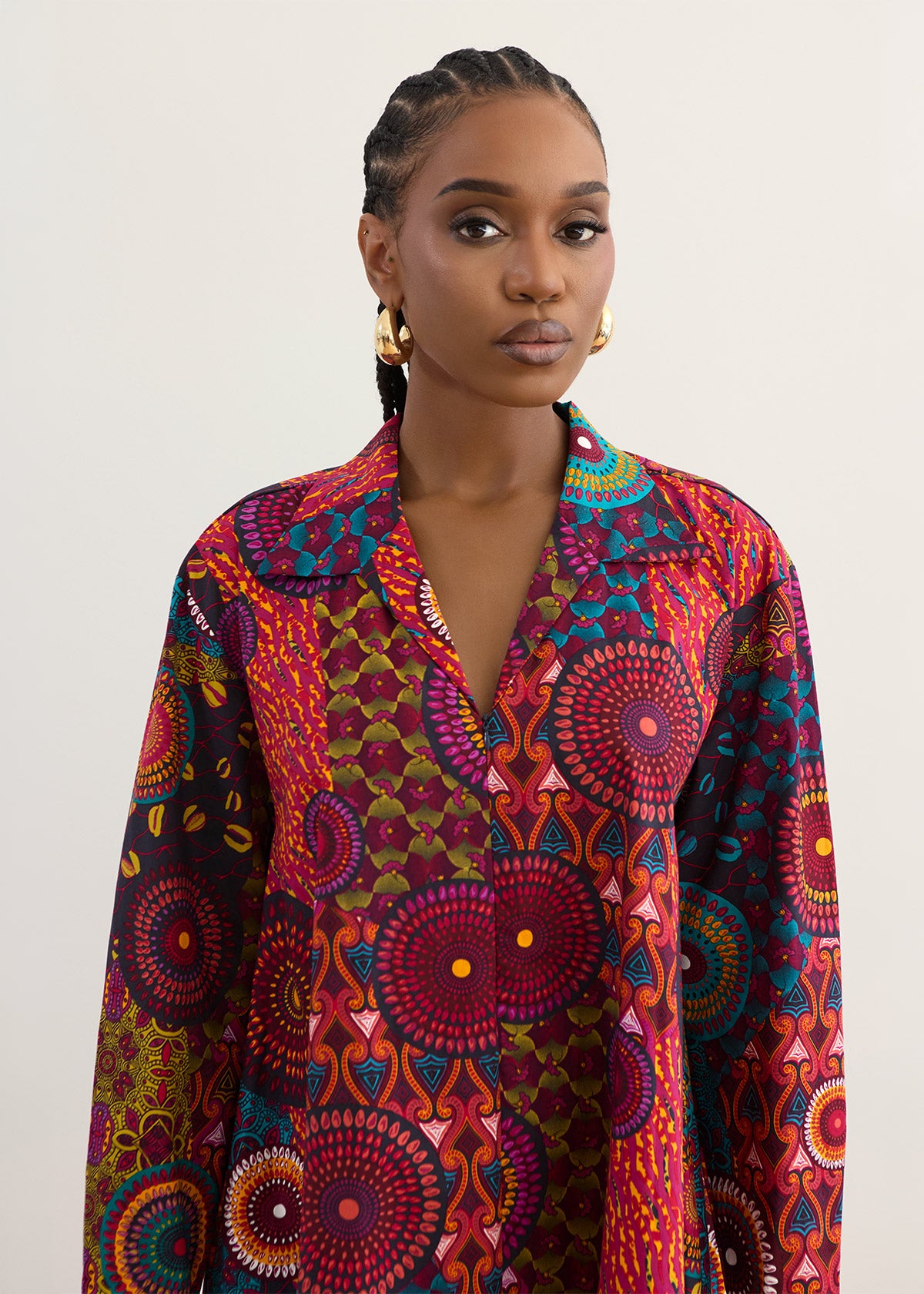 A woman wearing a long-sleeve button-up shirt with a vibrant, multicolored African wax print featuring circular mandala patterns in red, purple, orange, green, and blue, set against a plain light gray background.