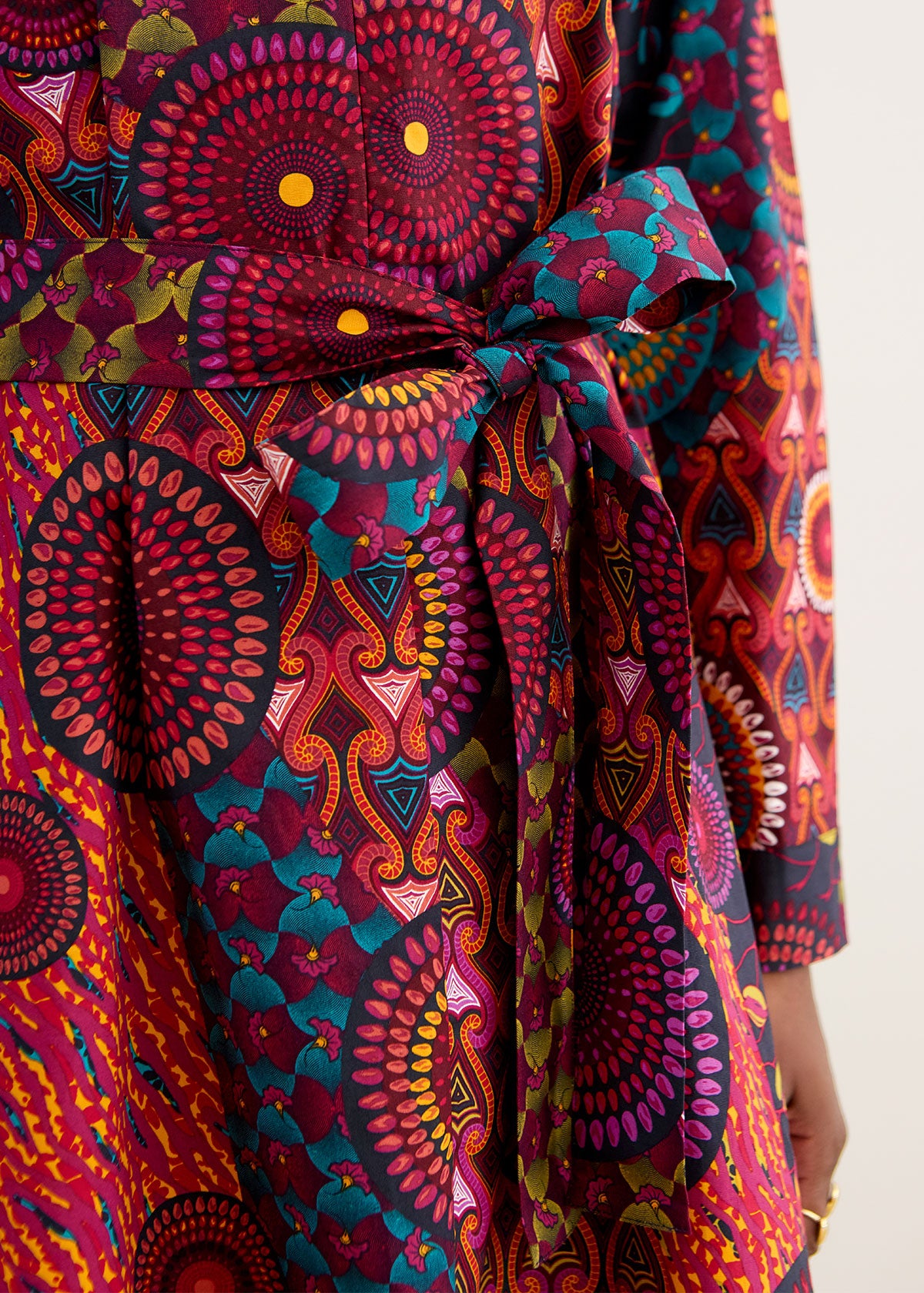 A close-up of a long-sleeved wrap dress featuring a vibrant, multicolored African wax print with circular mandala patterns and a matching self-tie belt at the waist.