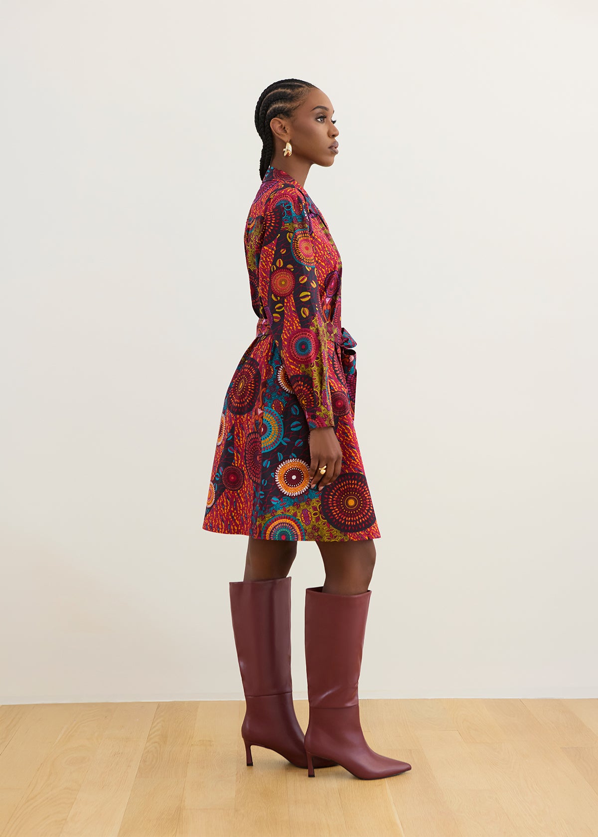 A model is shown from the side profile wearing a vibrant, multicolored patterned wrap dress with red, orange, and blue tones, paired with knee-high burgundy leather boots, set against a minimalist cream wall and light wood floor.