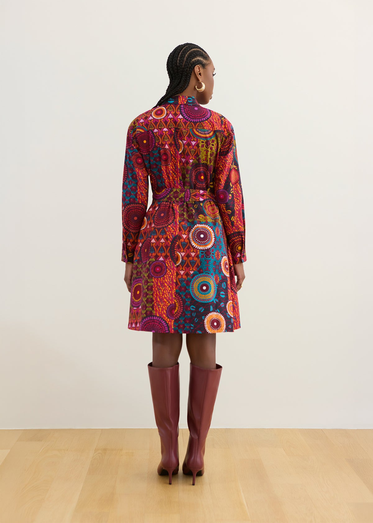 A model viewed from the back wearing a long-sleeve belted coat with a vibrant, multicolored circular mandala pattern, paired with knee-high burgundy boots, set against a minimalist beige wall and light wood floor.