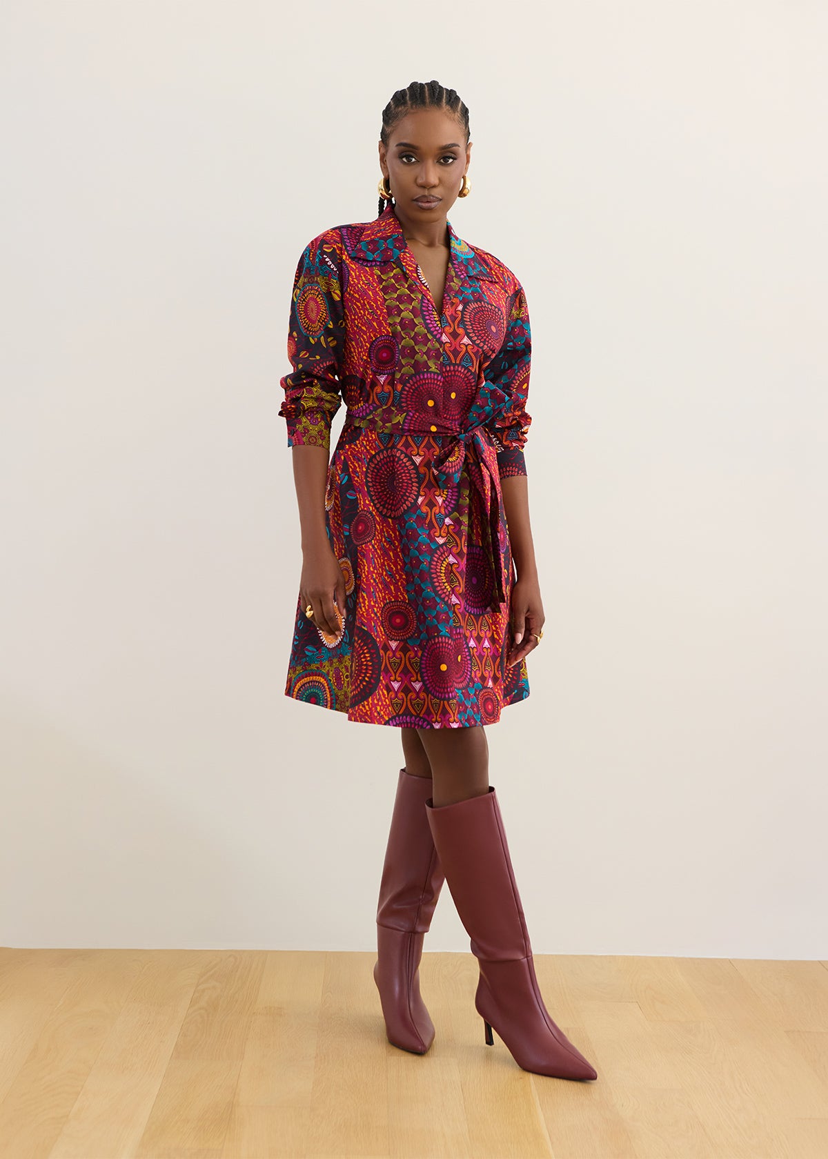A woman modeling a short wrap-style dress with a vibrant red, orange, and blue circular pattern, paired with burgundy knee-high heeled boots, standing against a plain off-white wall and light wood floor.