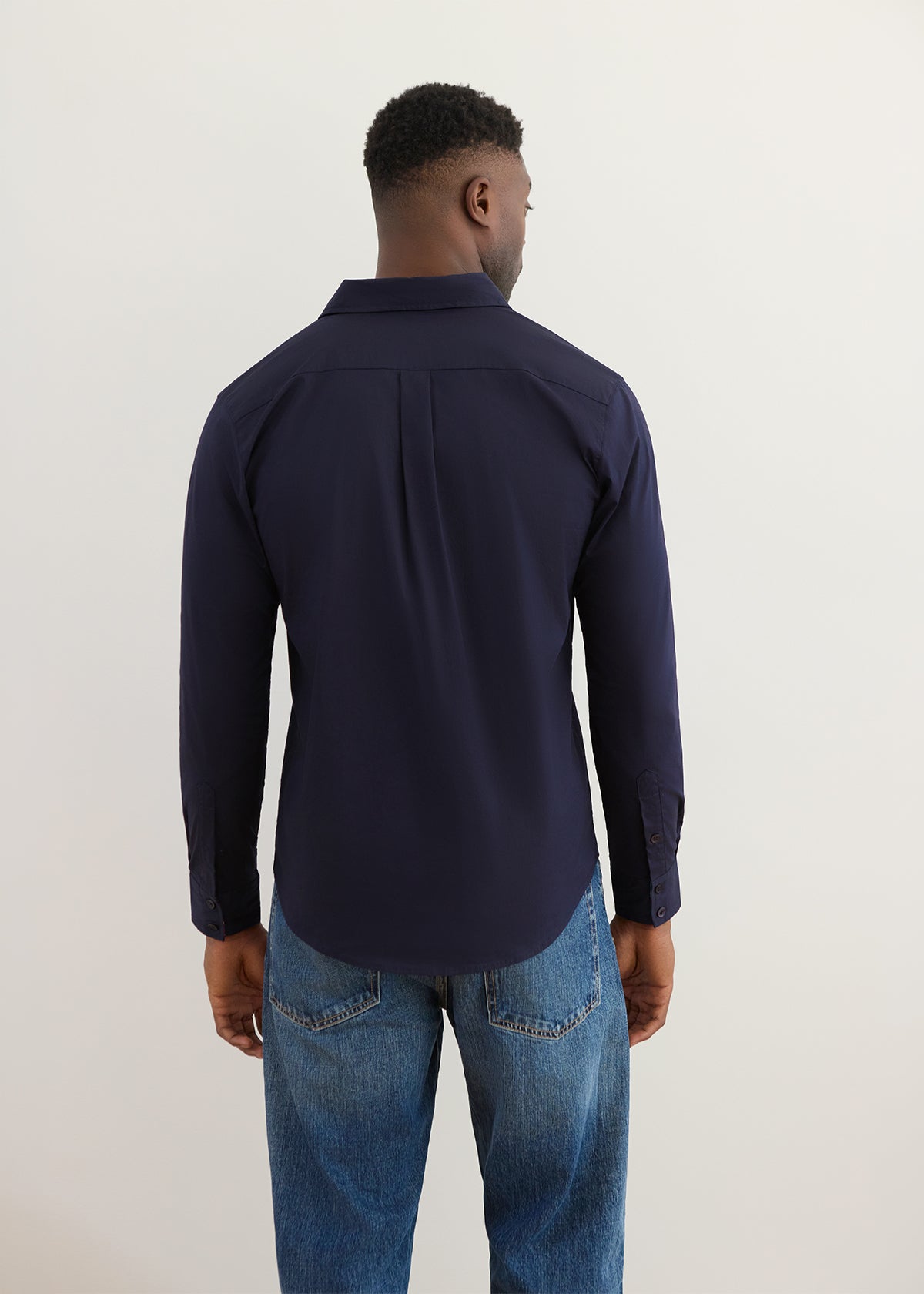 A man viewed from the back wearing a dark navy long-sleeve button-up shirt and blue jeans, standing against a plain light gray studio background.