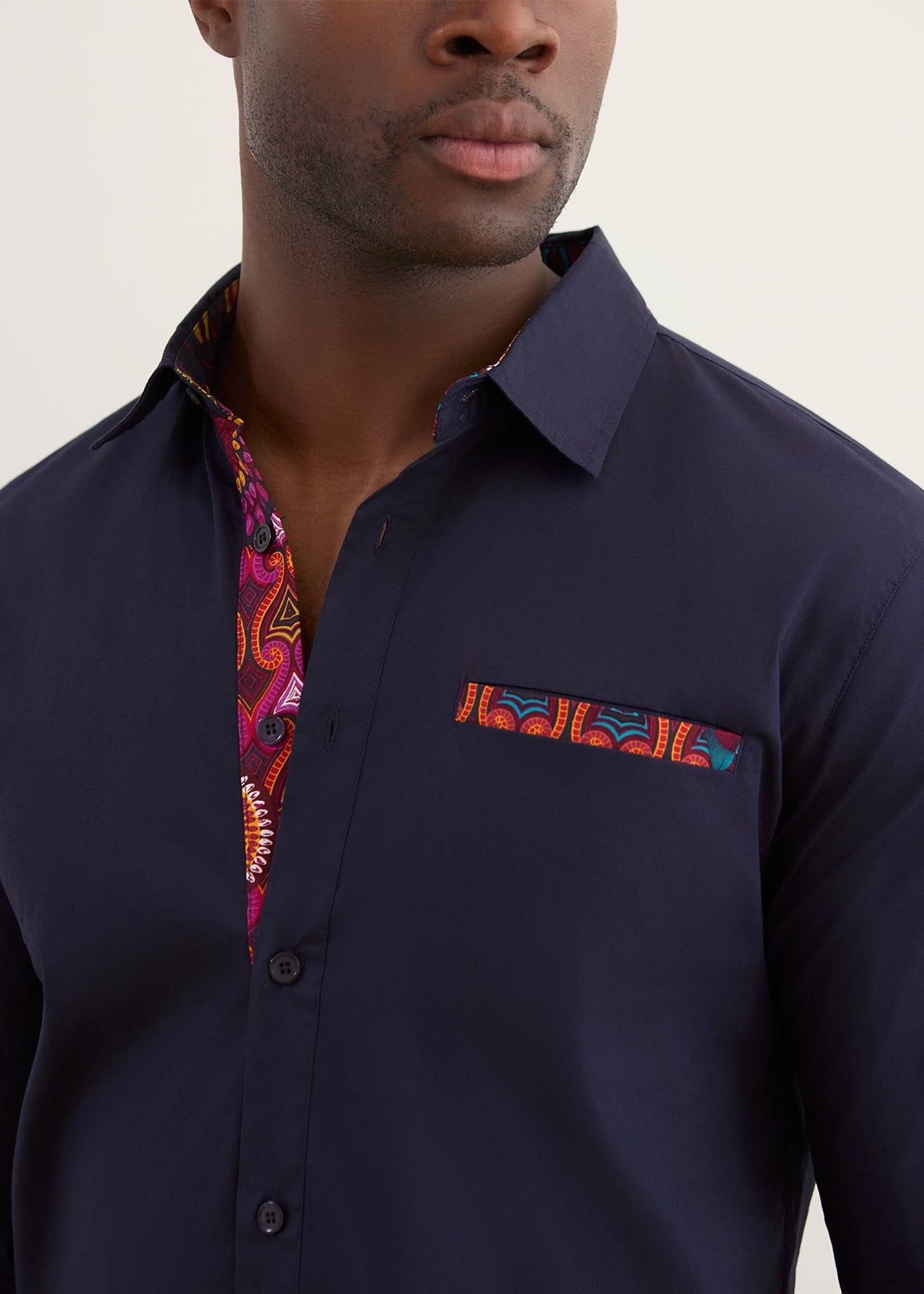 A close-up of a man wearing a dark navy blue button-down shirt with a vibrant, multicolored paisley pattern lining the inner collar and a matching patterned pocket square on the chest.