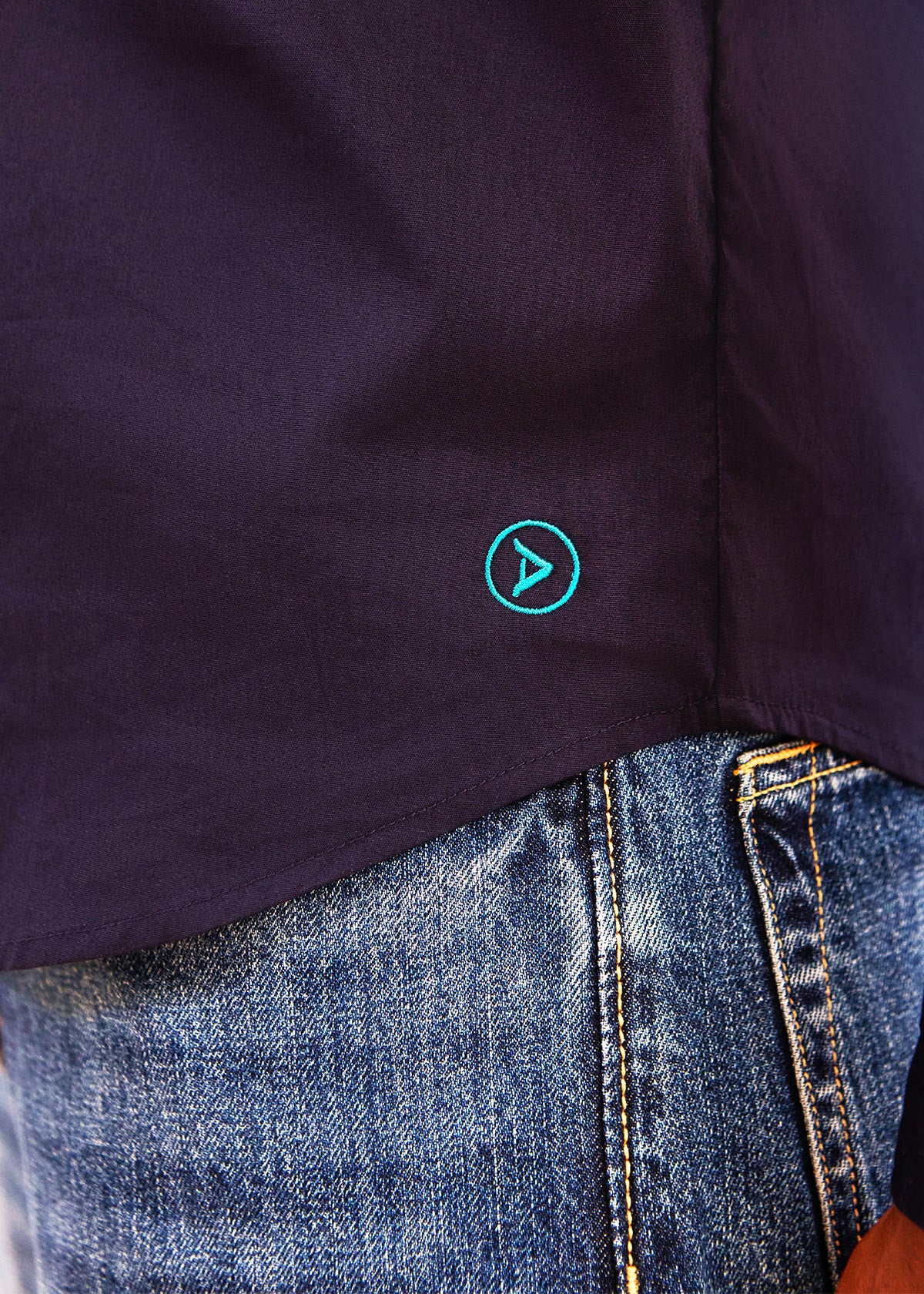 Close-up of a dark navy or black short-sleeve top worn with blue denim jeans, featuring a small teal circular logo with a play icon on the side.