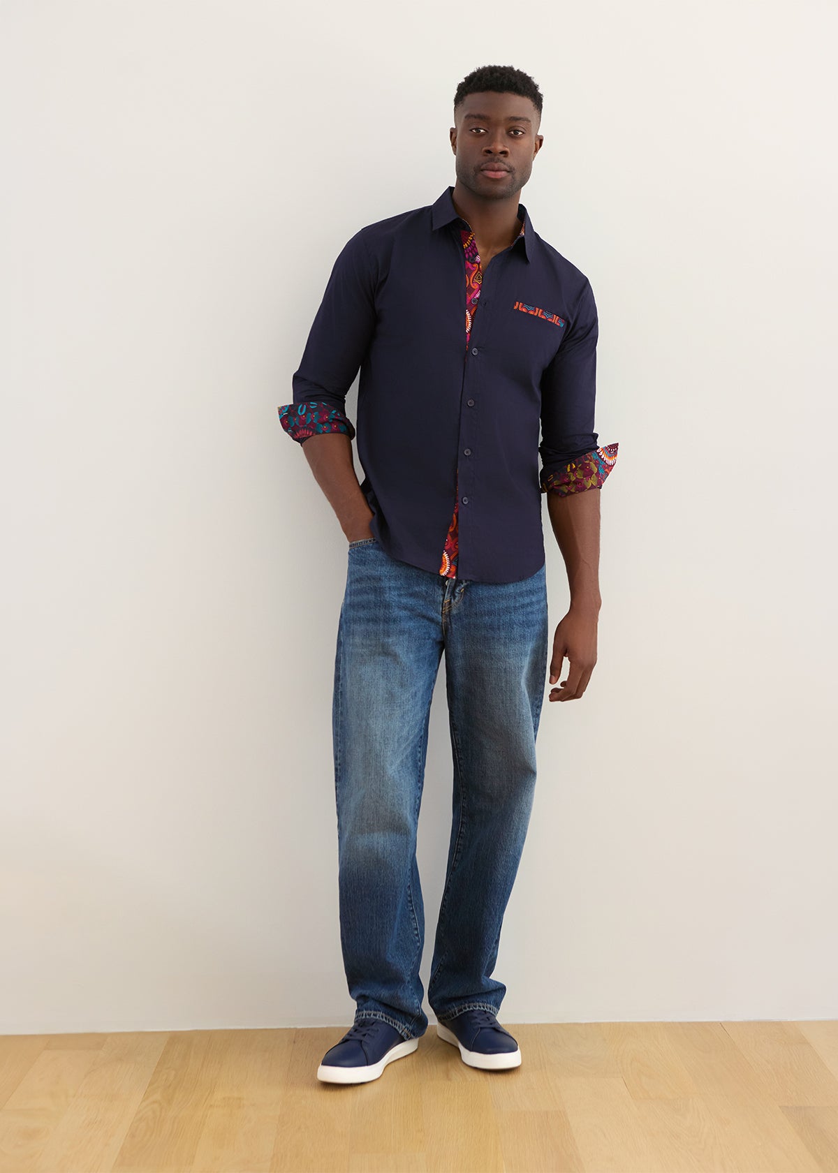 A man modeling a dark navy blue button-down shirt with colorful patterned cuffs and collar, paired with blue jeans and navy sneakers, standing against a plain off-white wall.