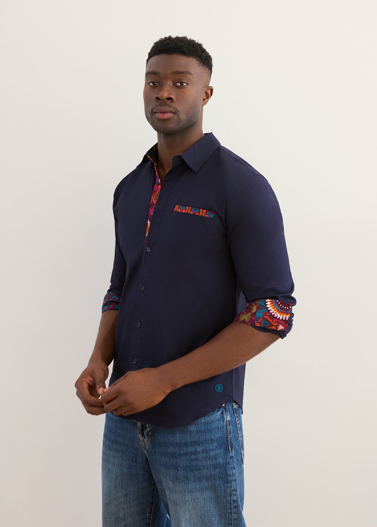 A man wearing a navy blue button-down shirt with colorful patterned inner placket and rolled-up sleeves, paired with blue jeans, standing against a plain light background.