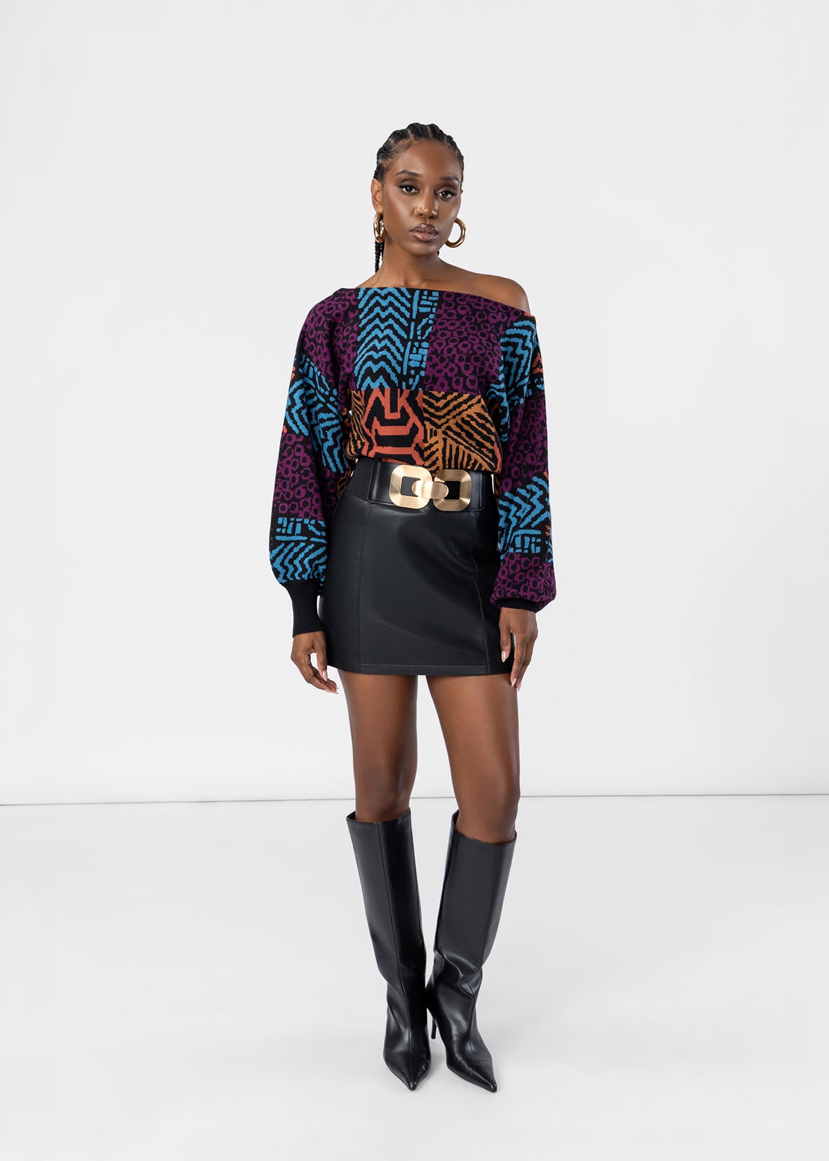 A model stands against a white studio background wearing a colorful off-the-shoulder long-sleeve top with geometric and leopard-like patterns in purple, blue, orange, and black, paired with a black mini skirt, a gold-tone belt, and black knee-high boots.
