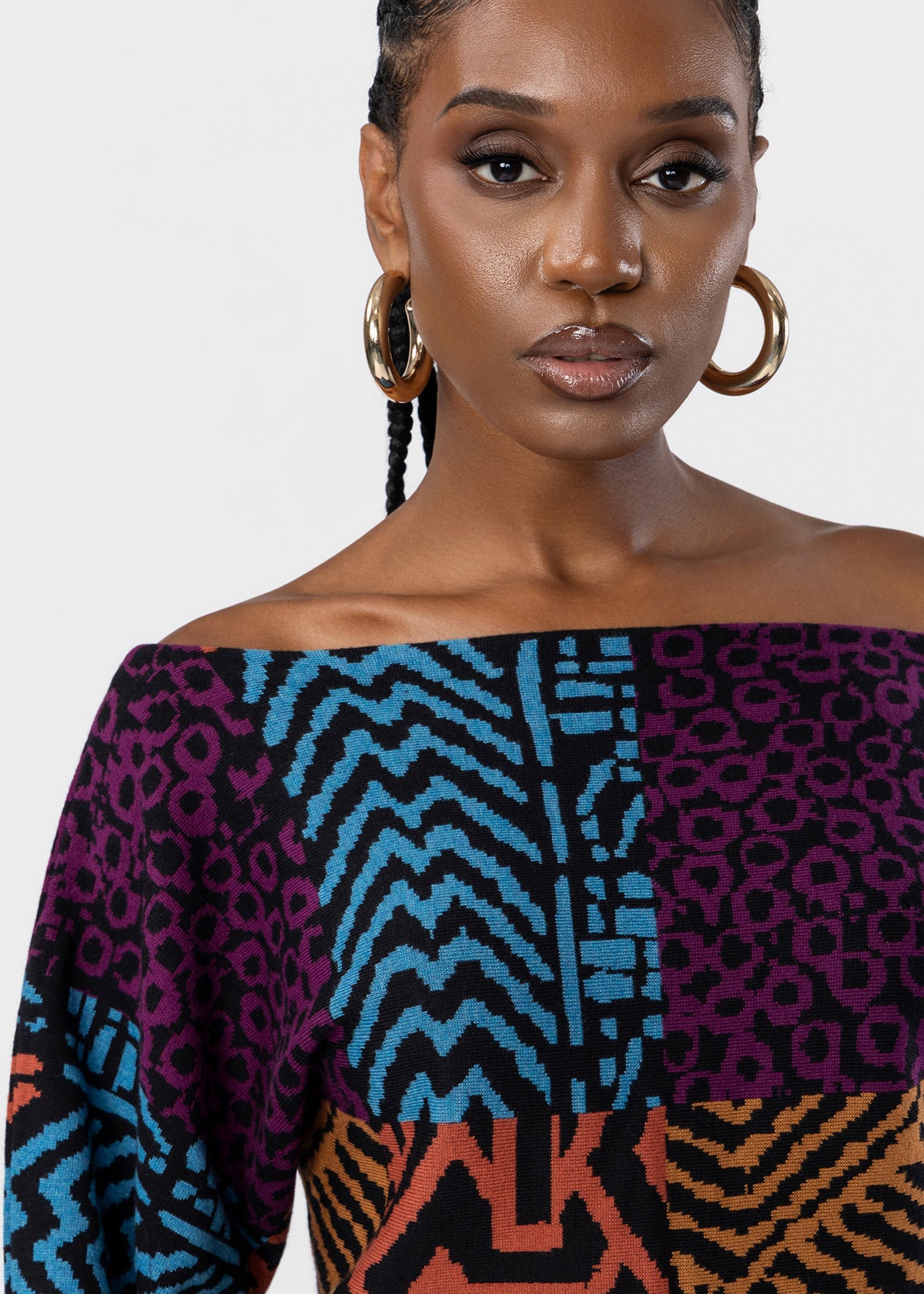 A close-up of a model wearing a vibrant off-the-shoulder knit top with a multicolor geometric and animal print patchwork design, paired with large gold hoop earrings against a light gray background.