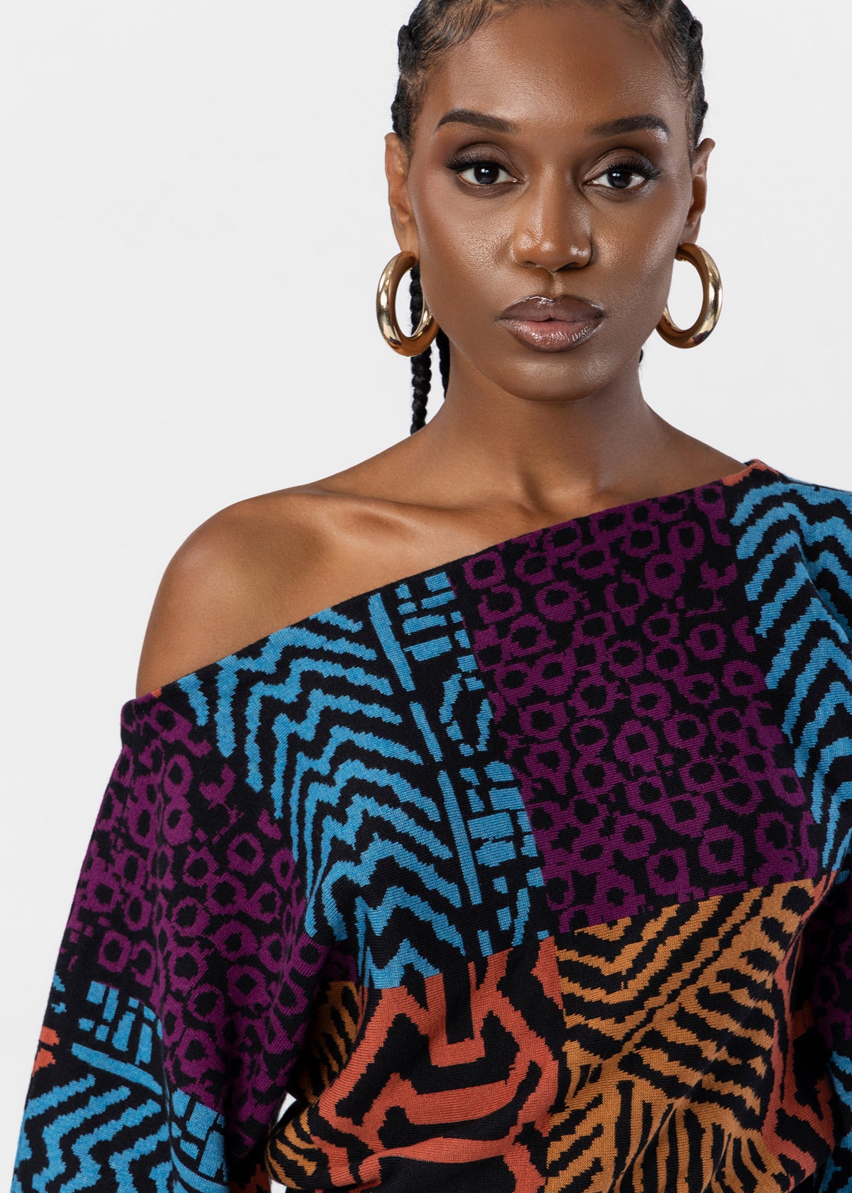 A close-up of a model wearing a vibrant, multi-colored off-the-shoulder top with a patchwork animal and geometric print. The top features panels of purple chevron, blue leopard, and orange zebra patterns on a black base.