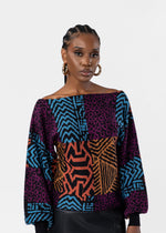 A woman modeling a colorful off-the-shoulder long-sleeve top with a patchwork print of purple leopard spots, blue chevron patterns, and orange geometric designs, set against a plain light gray background.