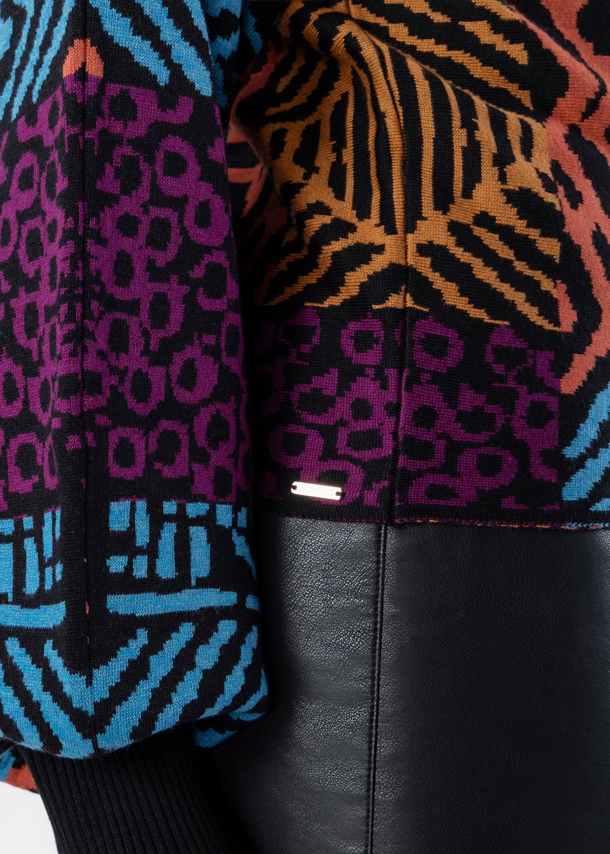 Close-up of a patterned knit top with black, purple, orange, and blue geometric and animal-inspired motifs, paired with black faux-leather bottoms.
