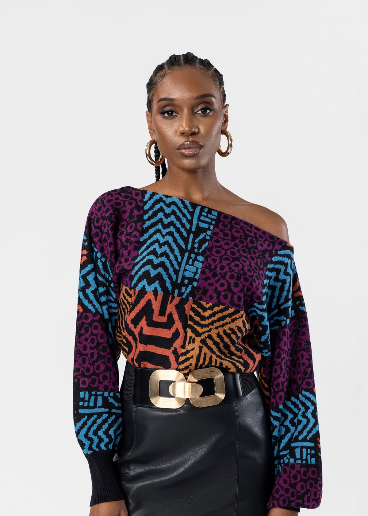 A model wearing a multicolor off-the-shoulder top with geometric and animal print panels in purple, blue, orange, and black, styled with a black leather skirt and a gold belt against a light gray background.