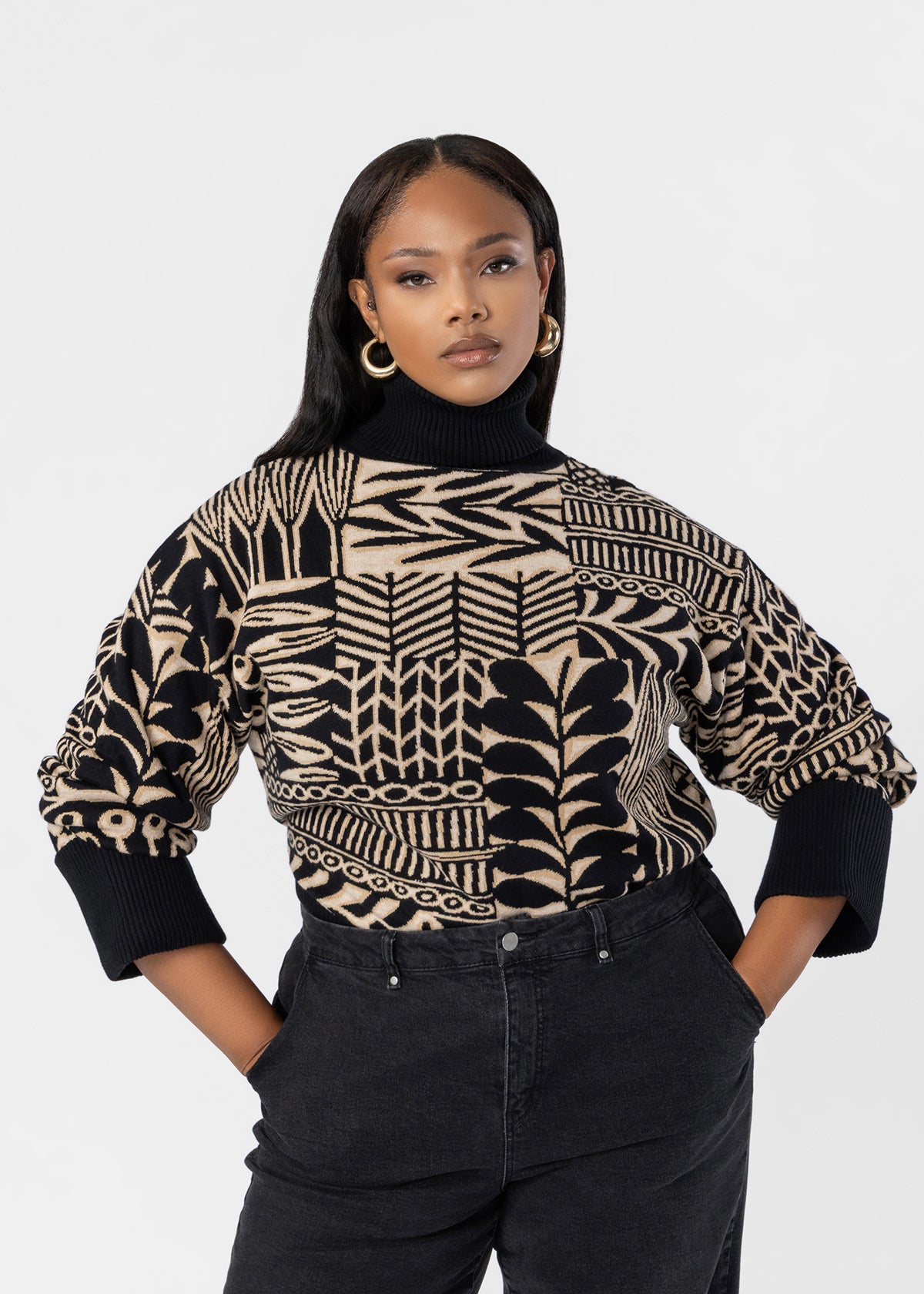 A woman modeling a black and beige geometric patterned knit turtleneck sweater with black pants, set against a plain light gray studio background.
