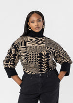 A woman modeling a black and beige geometric patterned knit turtleneck sweater with black pants, set against a plain light gray studio background.