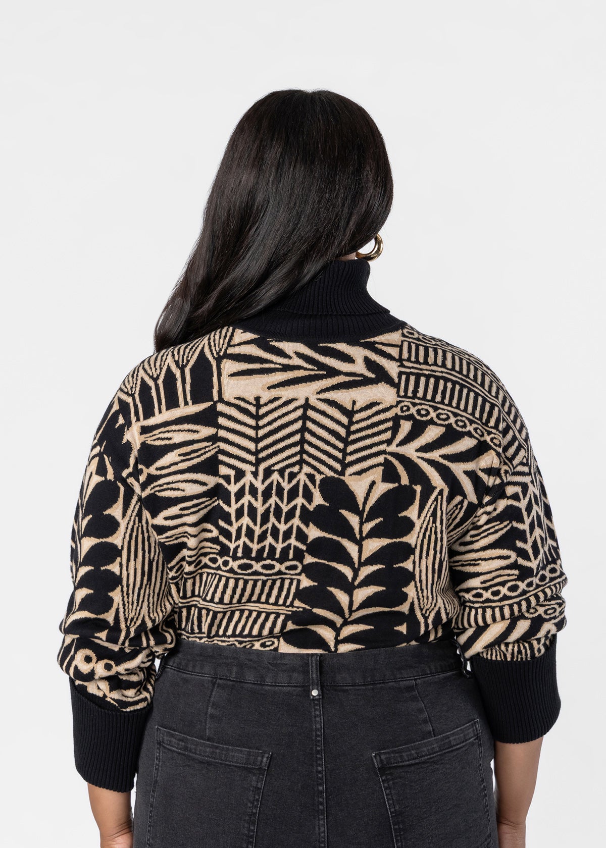 A woman viewed from behind wearing a black and beige patterned knit turtleneck sweater and dark gray jeans against a plain light gray studio background.