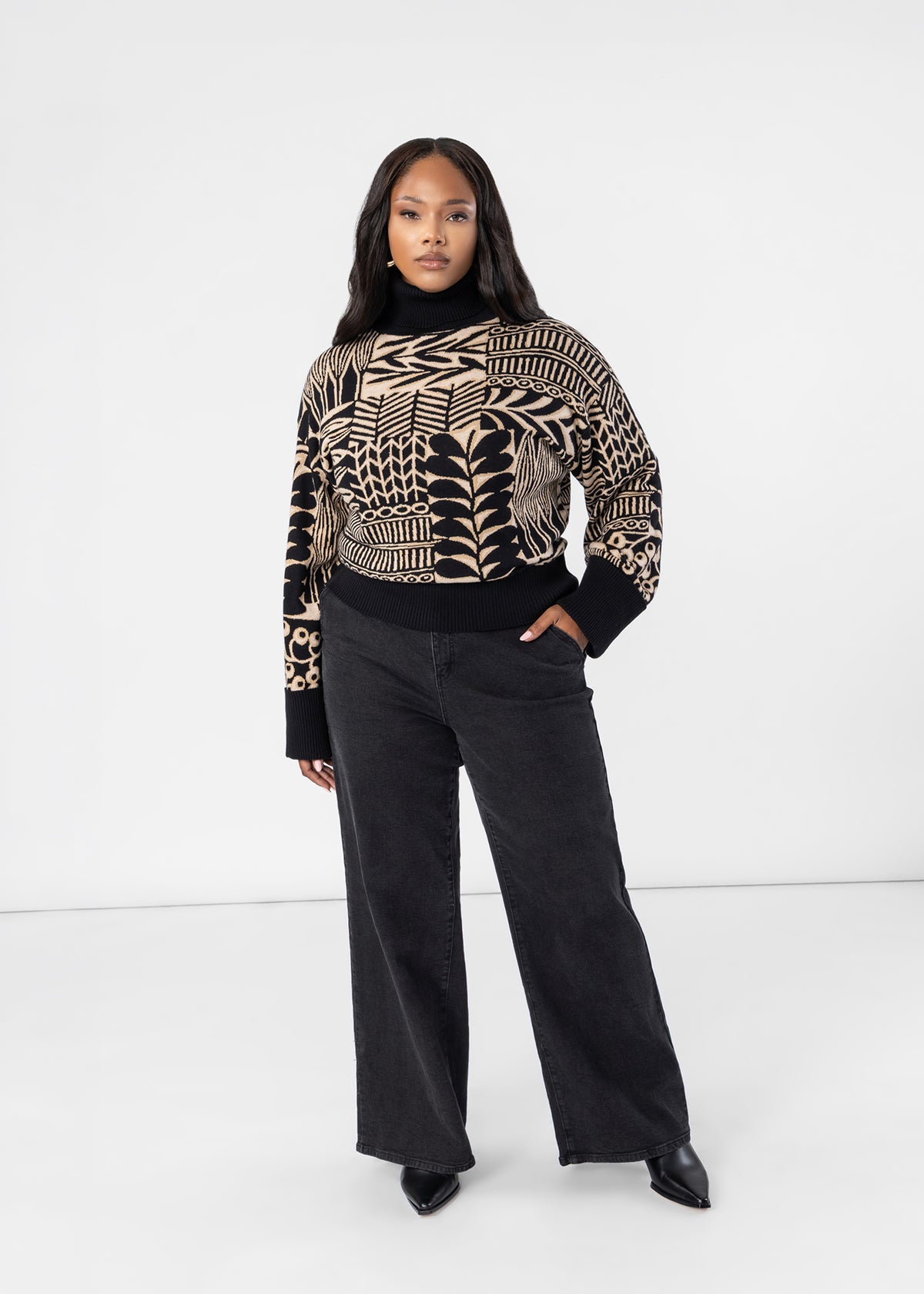 A woman modeling a black and beige patterned knit sweater with wide-leg black jeans, standing against a plain white studio background.