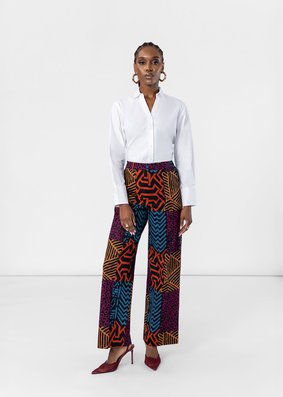 A model stands against a white studio background wearing a white long-sleeve button-up shirt, high-waisted wide-leg trousers with a vibrant multicolor geometric and tribal-inspired print in purple, orange, blue, and black, paired with burgundy pointed-toe heels.
