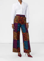 A model wearing a white button-down shirt and high-waisted, wide-leg trousers featuring a vibrant multicolor geometric and animal-print patchwork pattern in orange, blue, purple, and black. The model is also wearing burgundy pointed-toe heels against a clean white studio background.