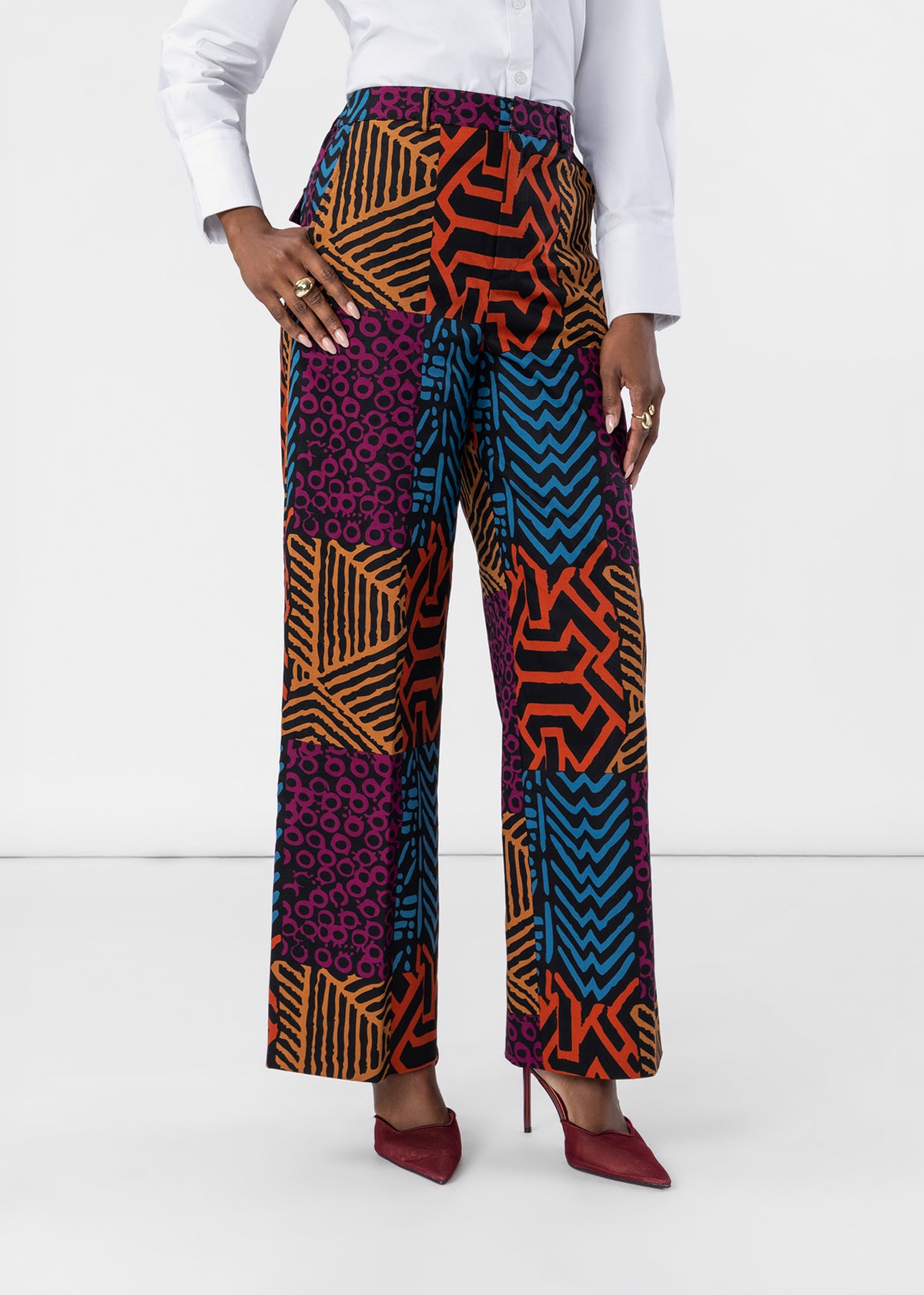 A model wearing high-waisted, wide-leg trousers with a vibrant multicolor patchwork print of geometric and circular patterns in orange, blue, purple, and black. The model is styled with a white long-sleeve shirt and burgundy pointed-toe heels against a plain white studio background.