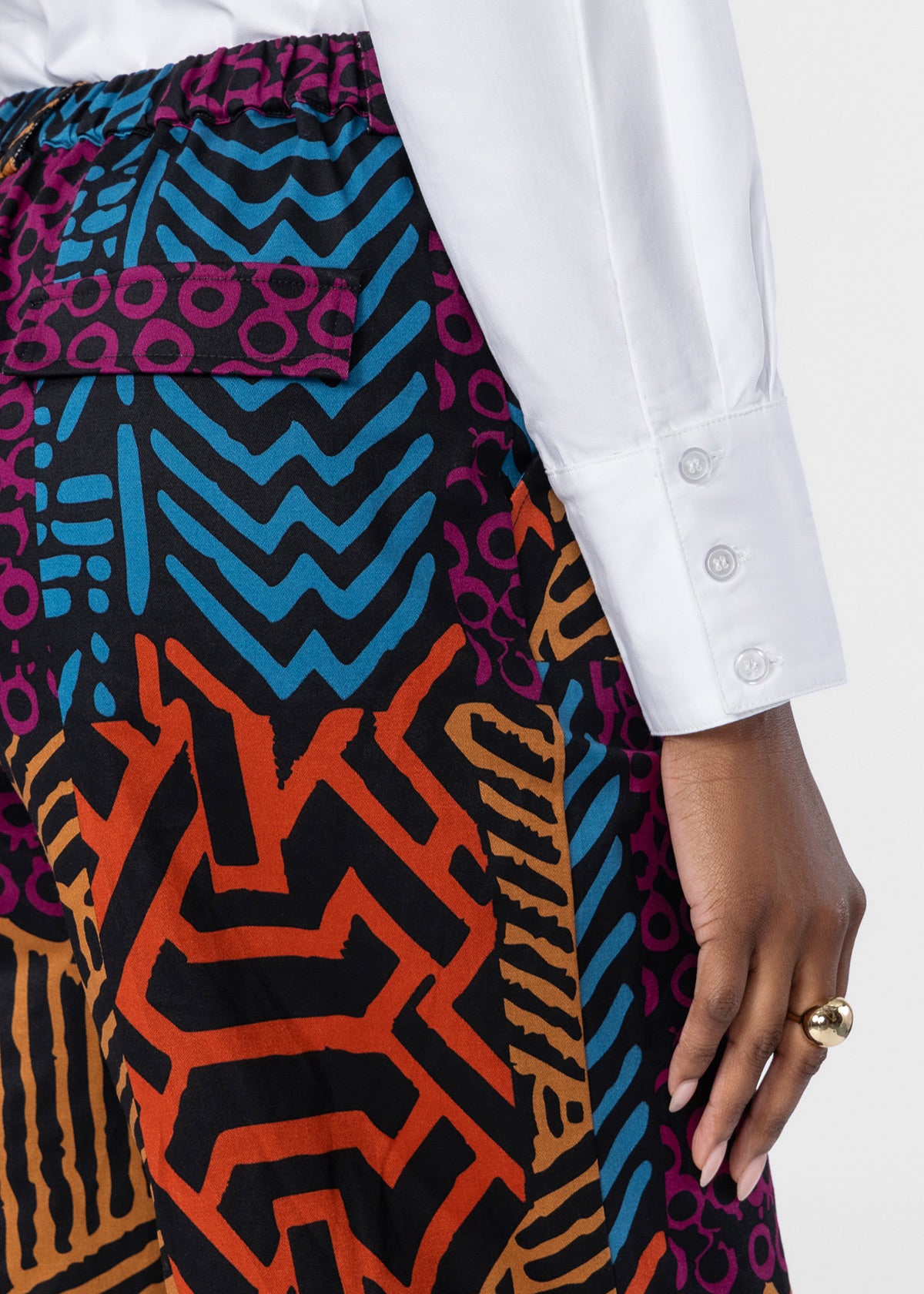 Close-up of a person wearing patterned wide-leg trousers with a white button-up shirt. The trousers feature a bold geometric and abstract print in black, orange, blue, purple, and tan.
