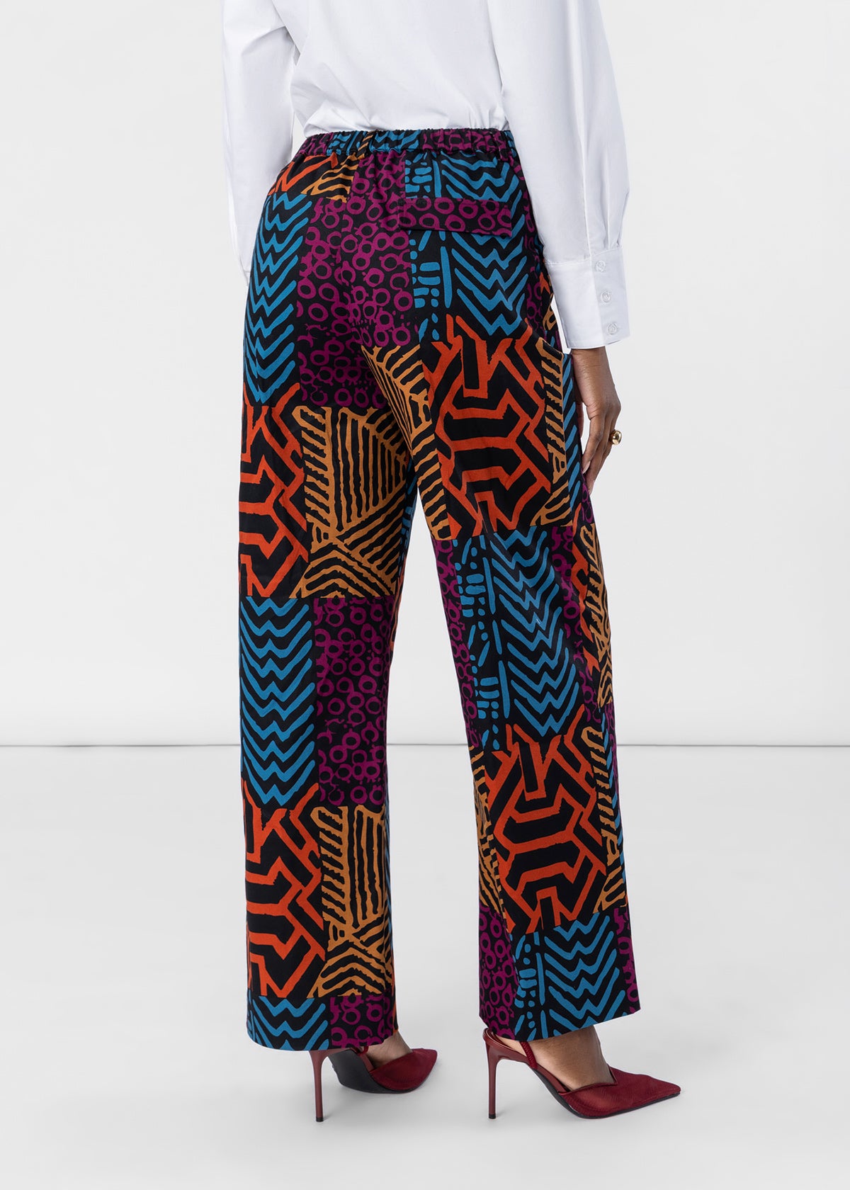 A person shown from the back wearing high-waisted, wide-leg trousers with a vibrant multicolor geometric and abstract patchwork print in orange, blue, purple, and black. The model is also wearing a white long-sleeve blouse and burgundy pointed-toe heels against a clean white studio background.