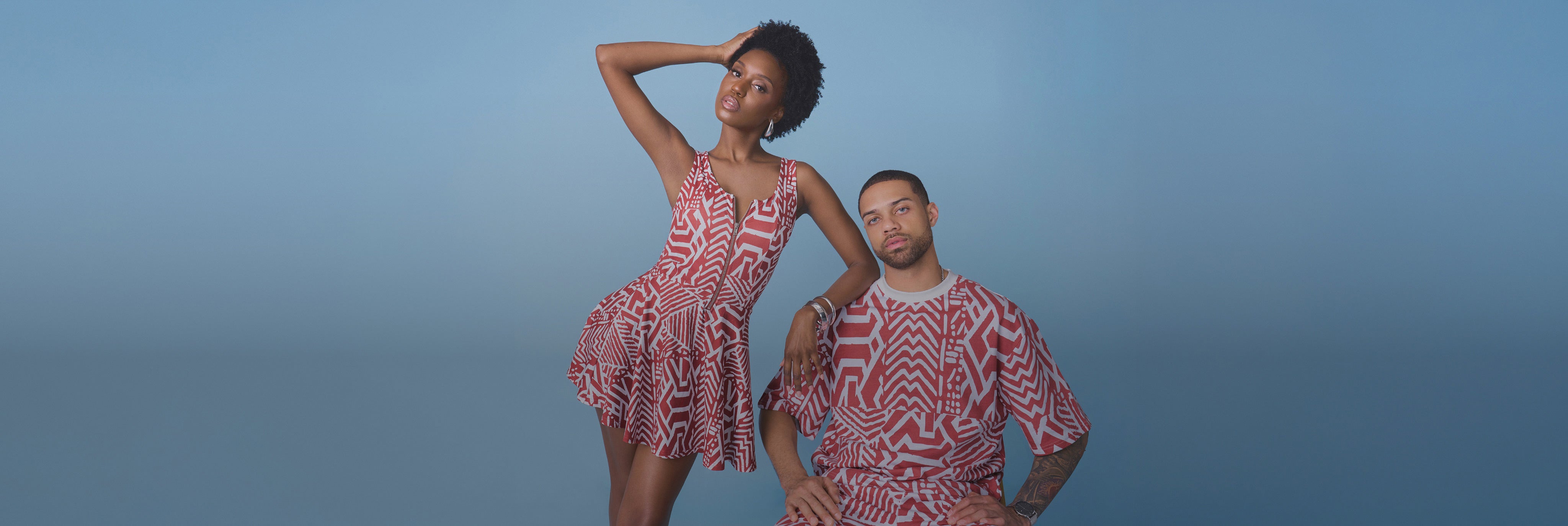 Two models posing in matching red-and-white African-inspired print outfits against a blue studio background
