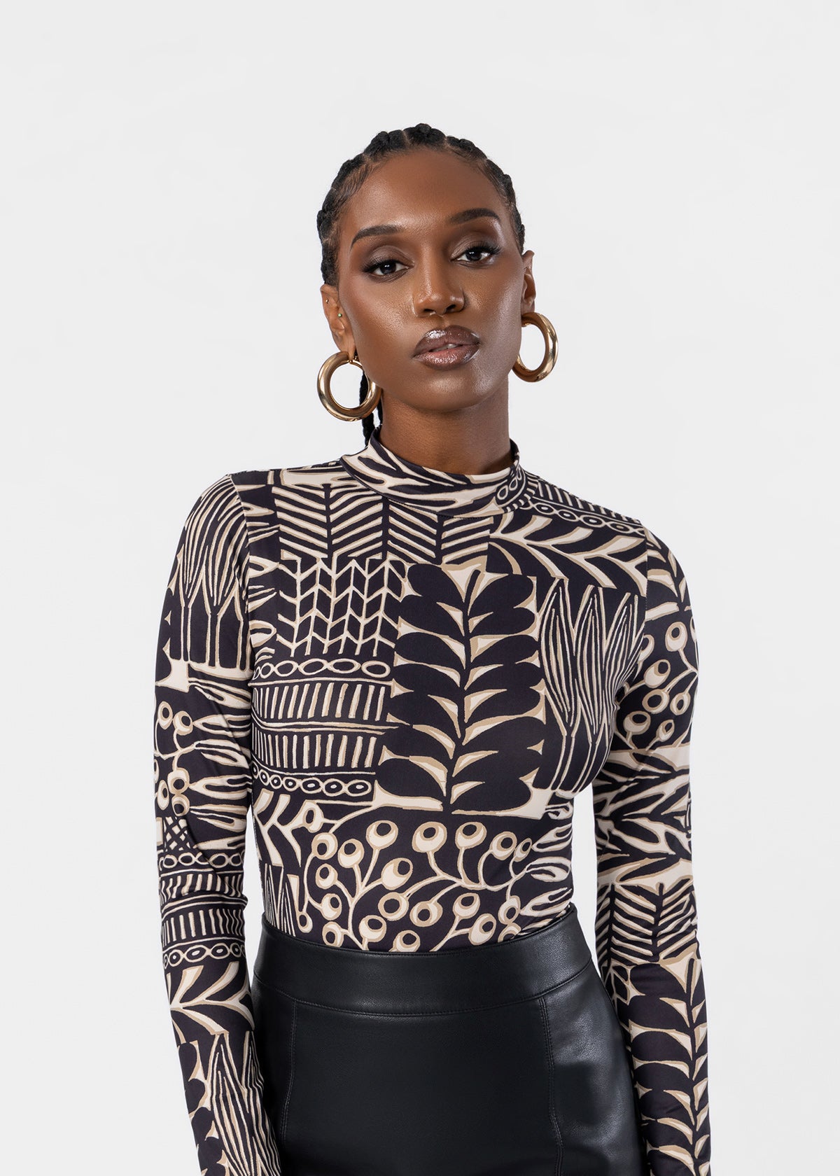A woman modeling a long-sleeve mock neck top with a black and beige botanical pattern, paired with black leather pants, against a light gray studio background.