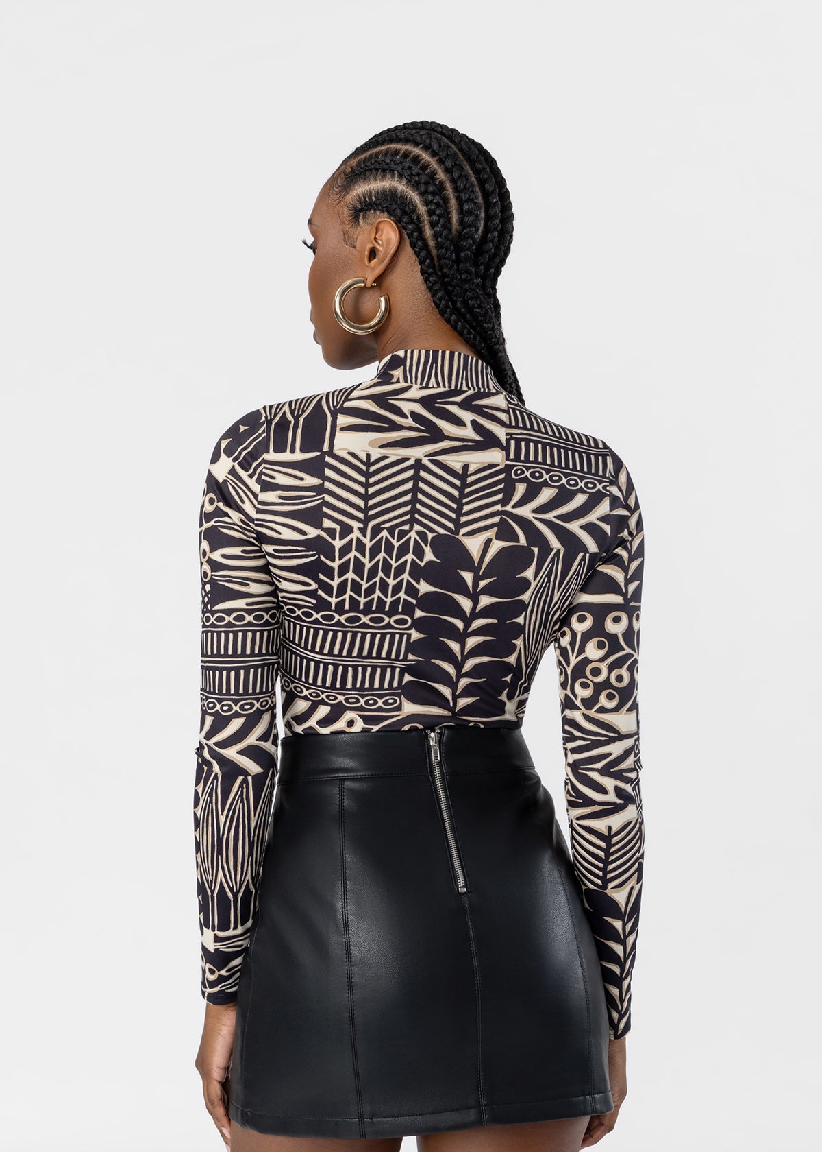 A woman shown from the back wearing a long-sleeve mock-neck top with a black and beige geometric and botanical pattern, paired with a black faux-leather mini skirt, against a light gray studio background.