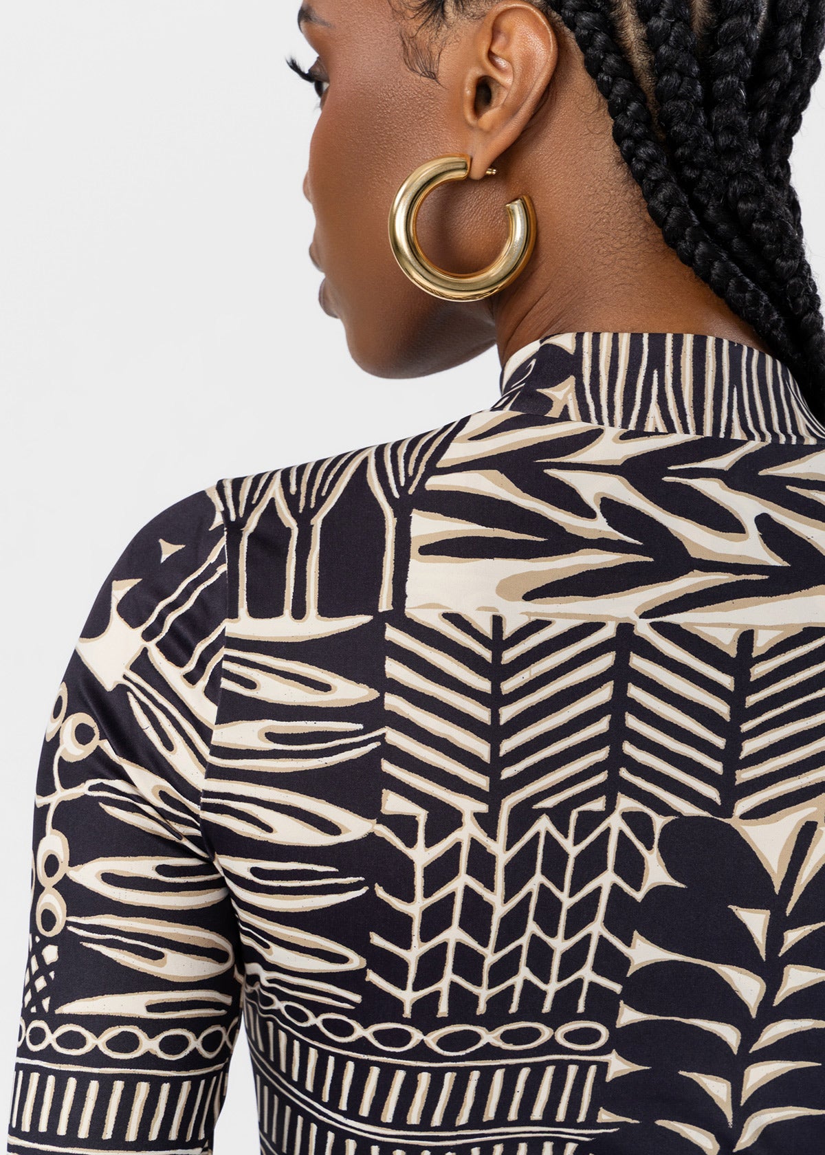 Close-up back view of a woman wearing a high-neck top with a bold black and cream botanical pattern, paired with large gold hoop earrings, against a light gray studio background.