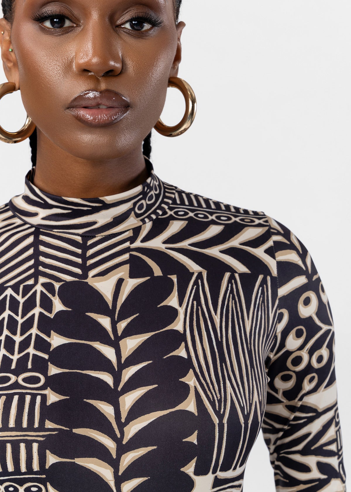 A close-up of a model wearing a long-sleeve mock neck top with a bold black and beige botanical block print pattern, paired with large gold hoop earrings against a light gray background.