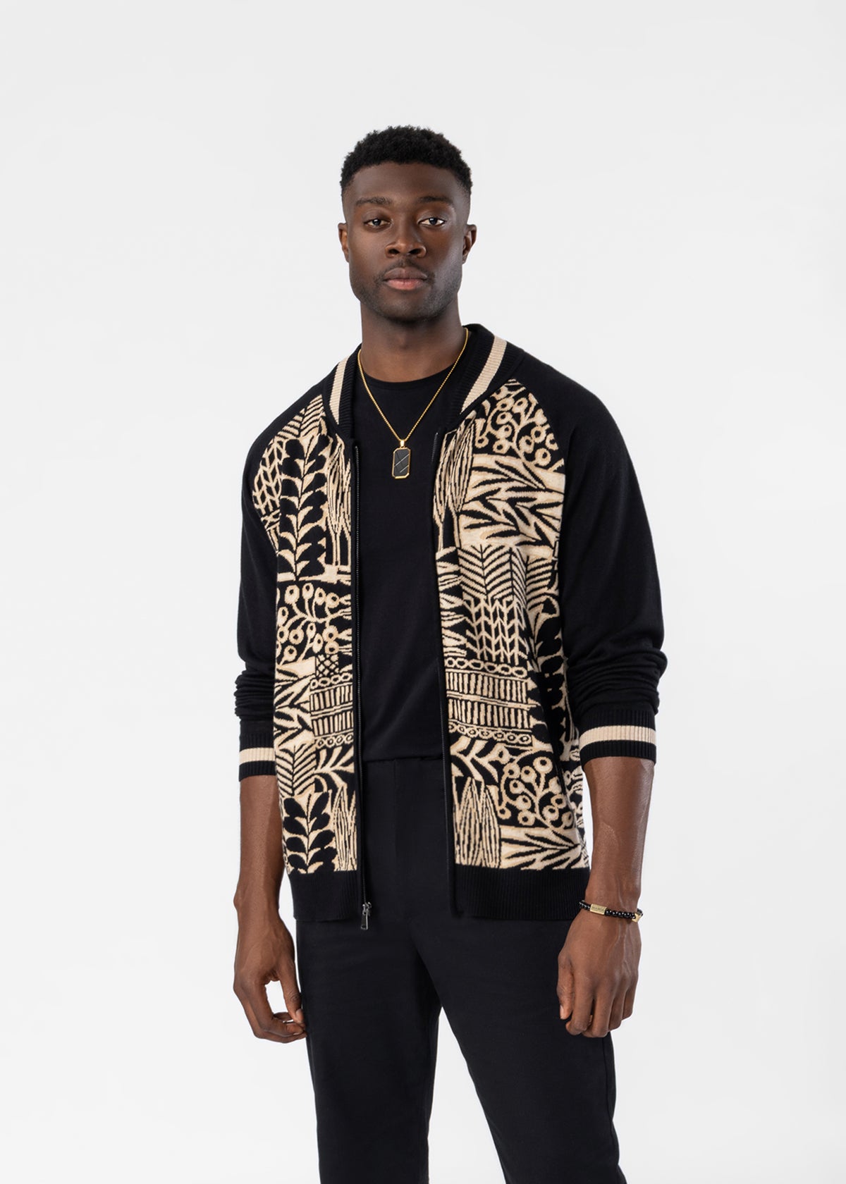 A man modeling a black and beige patterned knit cardigan over a black t-shirt and black pants, shot against a solid light gray background.