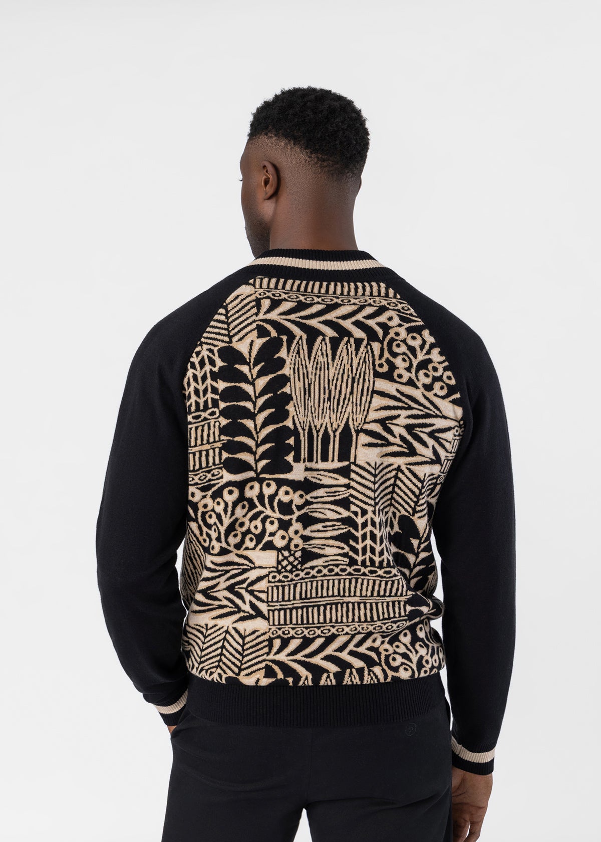 A man viewed from the back wearing a black and beige patterned knit sweater with solid black raglan sleeves and a detailed botanical design on the back.