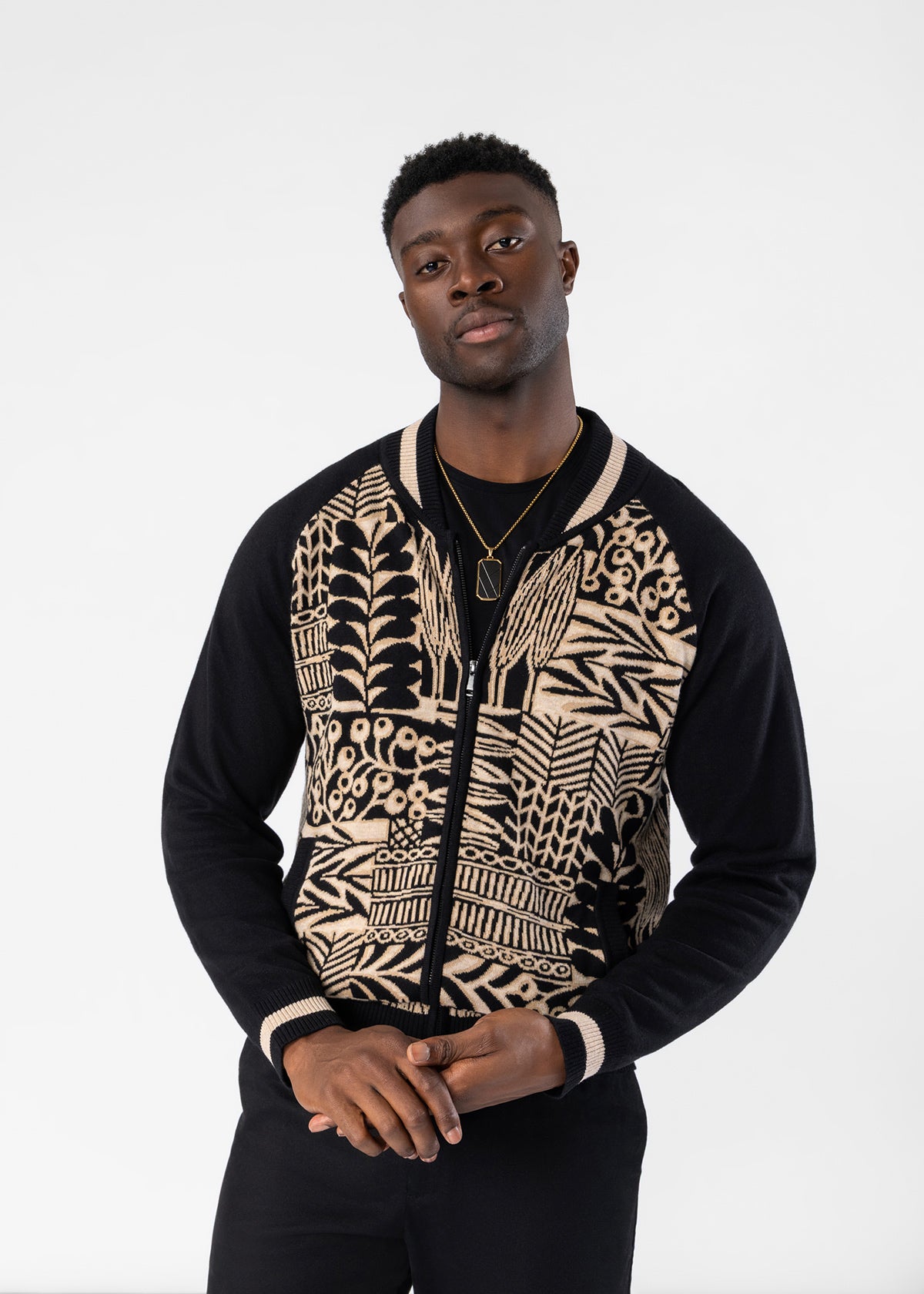 A man wearing a black and beige patterned bomber jacket with a black zipper, black sleeves, and beige ribbed trim on the collar, cuffs, and hem, standing against a plain white studio background.