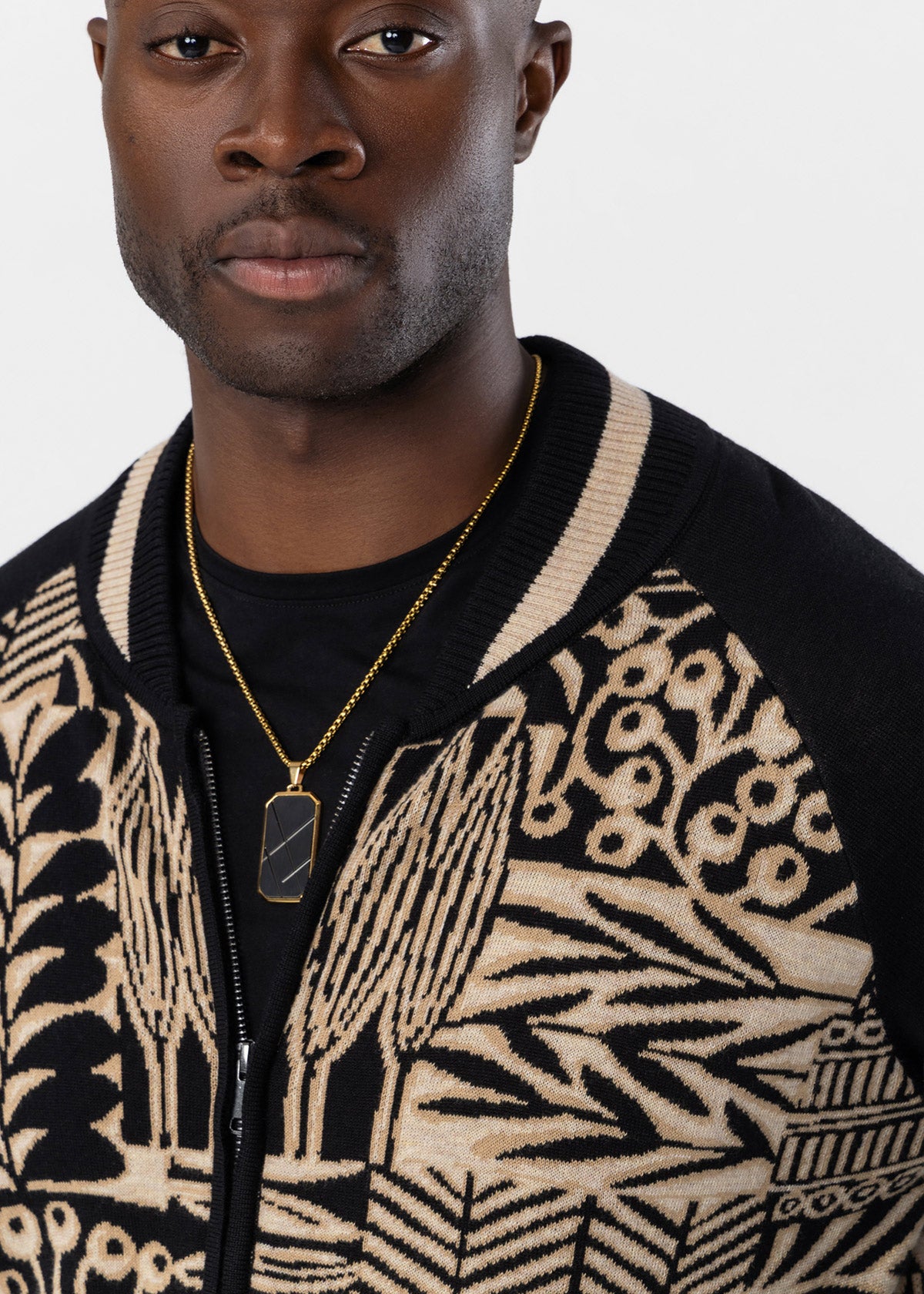 A close-up of a man wearing a black and beige patterned knit zip-up cardigan over a black t-shirt, accessorized with a gold chain necklace.