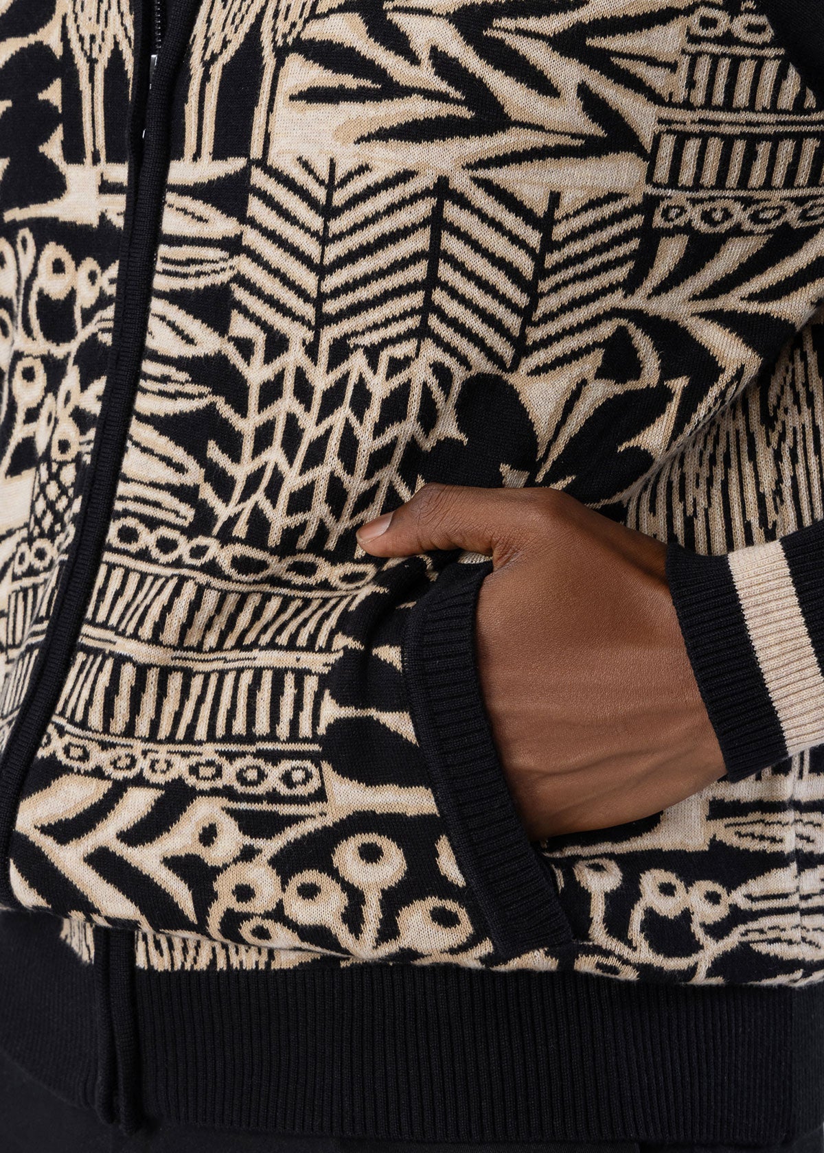 Close-up of a black and cream knit zip-up sweater with a hand in the pocket, showing detailed knit patterns and ribbed cuffs.