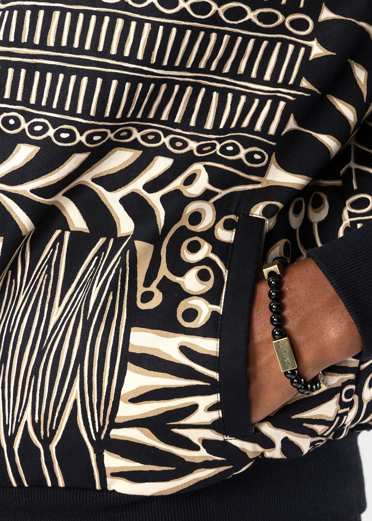 Close-up of a black and beige patterned knit garment with a side pocket, worn by a model whose hand and wrist with a black beaded bracelet are visible.