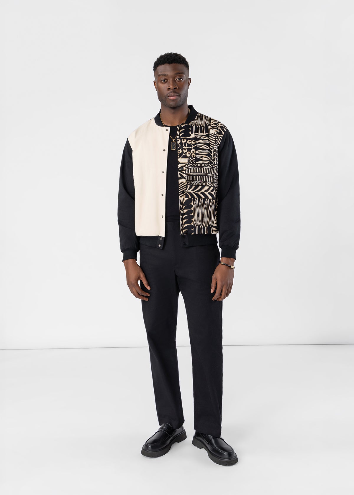 A man modeling a unique color-blocked bomber jacket with cream panels, black sleeves, and a black-and-beige geometric pattern across the chest, paired with black trousers and shoes, against a plain white studio background.